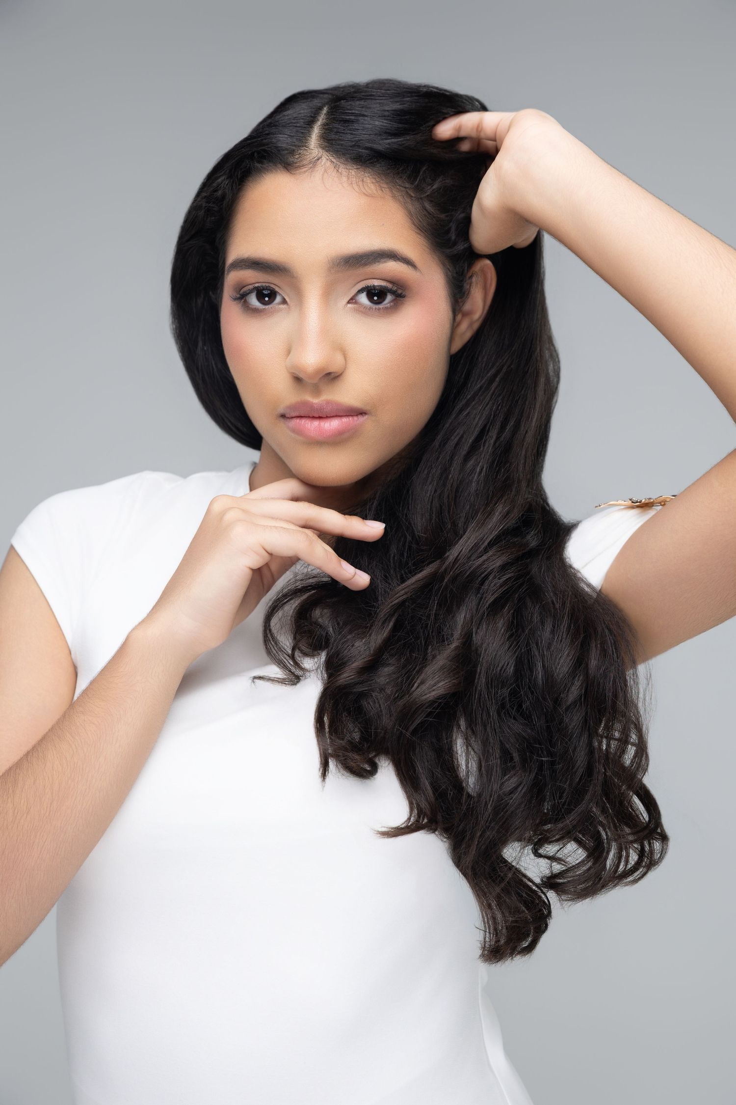 Young woman with long wavy dark hair poses in white t-shirt, hands in hair, against gray background.
