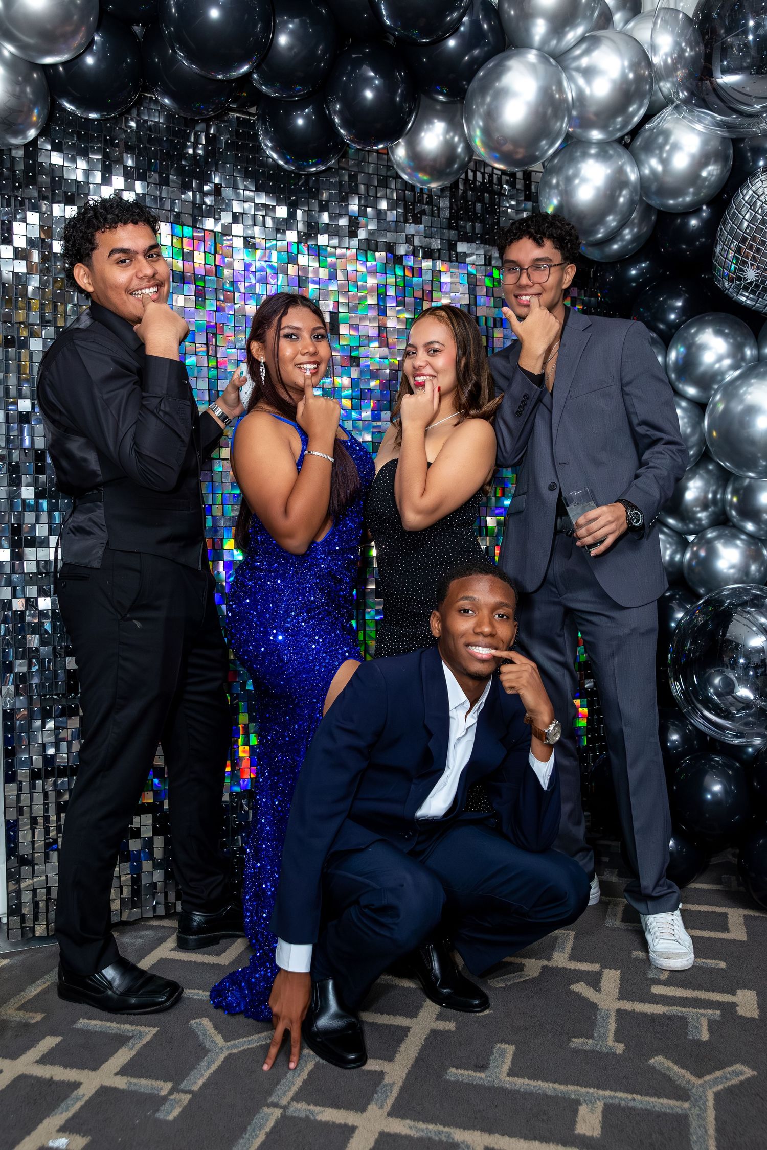 A group in formal attire poses together against a backdrop of silver balloons and sparkly decorations at an event.