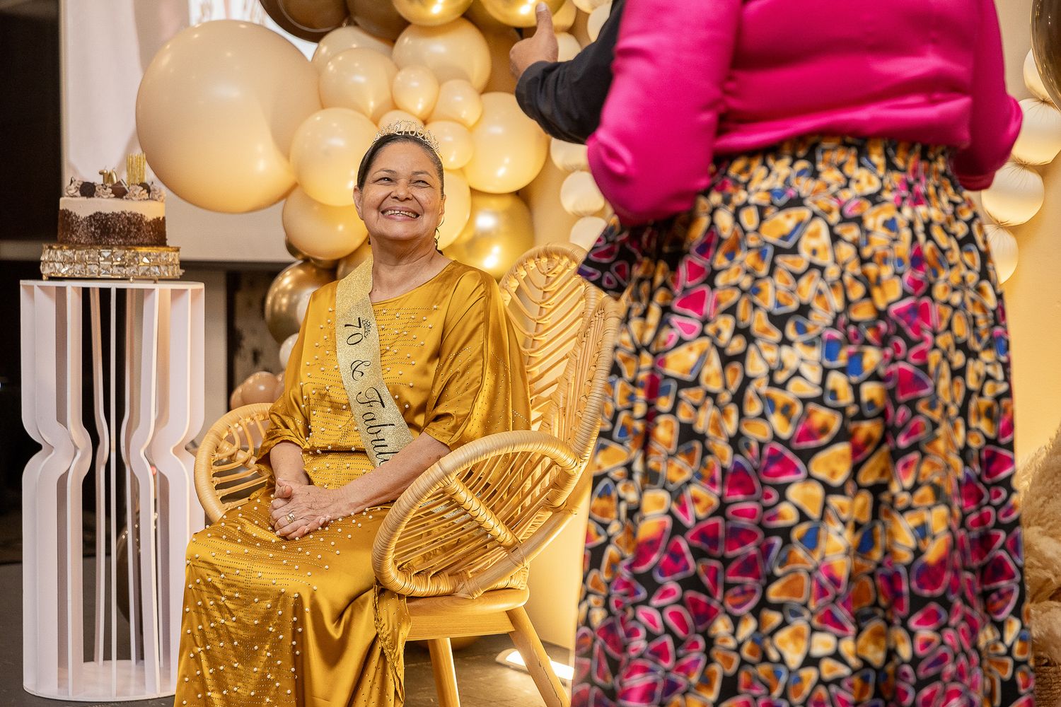Woman in golden gown sits in rattan chair, smiling at guest in floral skirt, surrounded by gold balloon decor.
