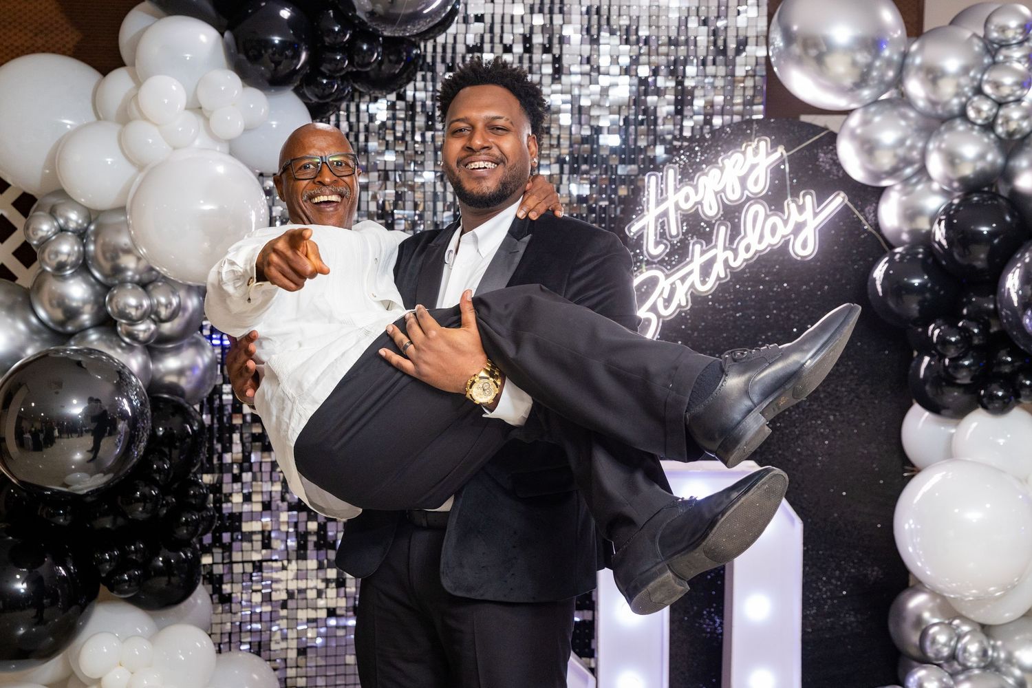 Two men celebrating a birthday party, one carrying the other, with black and silver balloon decorations.