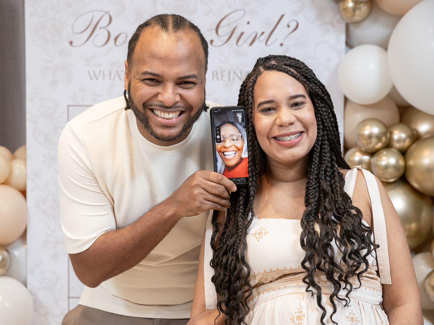 Couple smiling at gender reveal party, holding phone with video call, balloon backdrop with Boy or Girl sign.