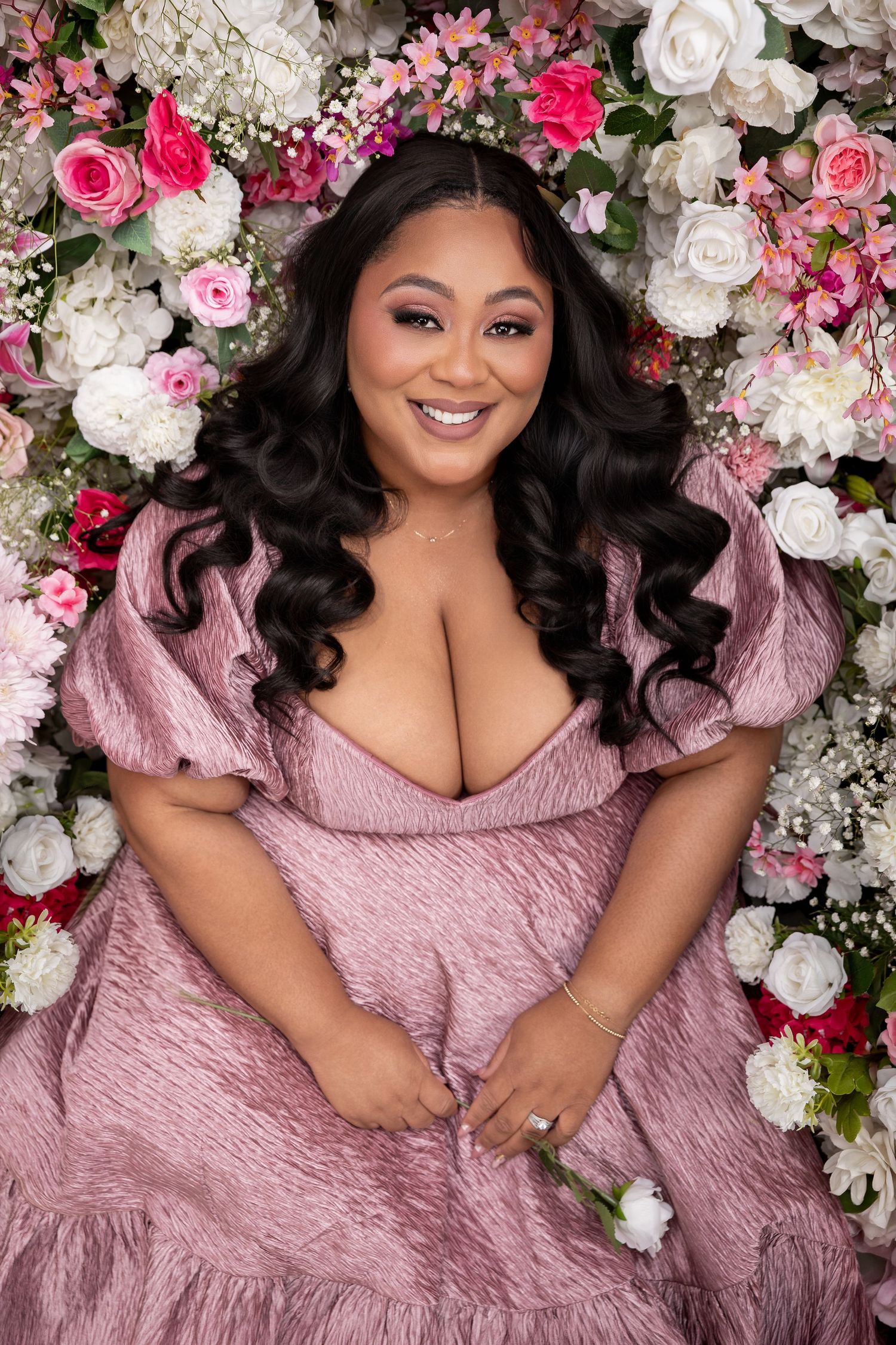 Woman in pink puff-sleeve dress smiling among white and pink roses in a floral portrait.