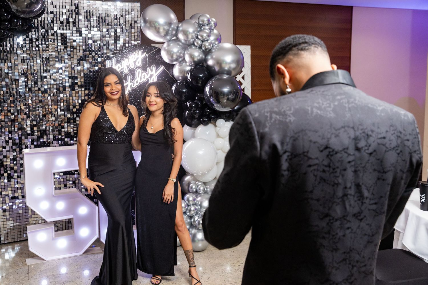 Two women in black dresses pose for photos at a birthday party with silver and black balloon decorations.