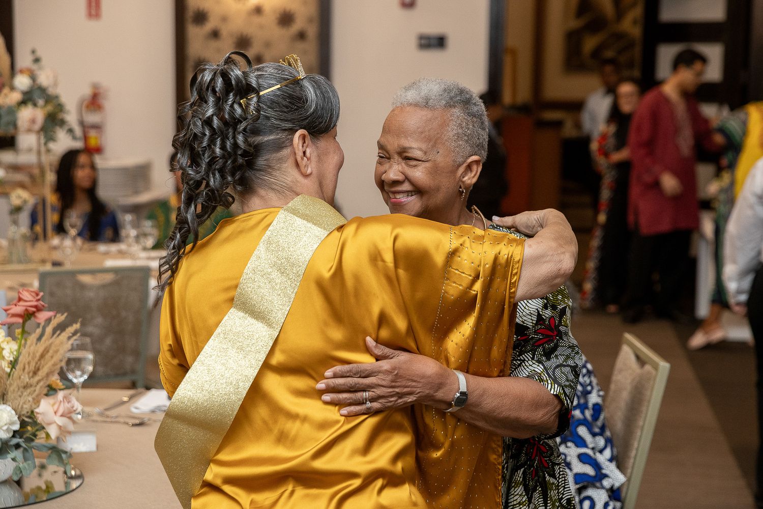 Two women in golden attire share a warm embrace at an elegant indoor celebration or wedding reception.