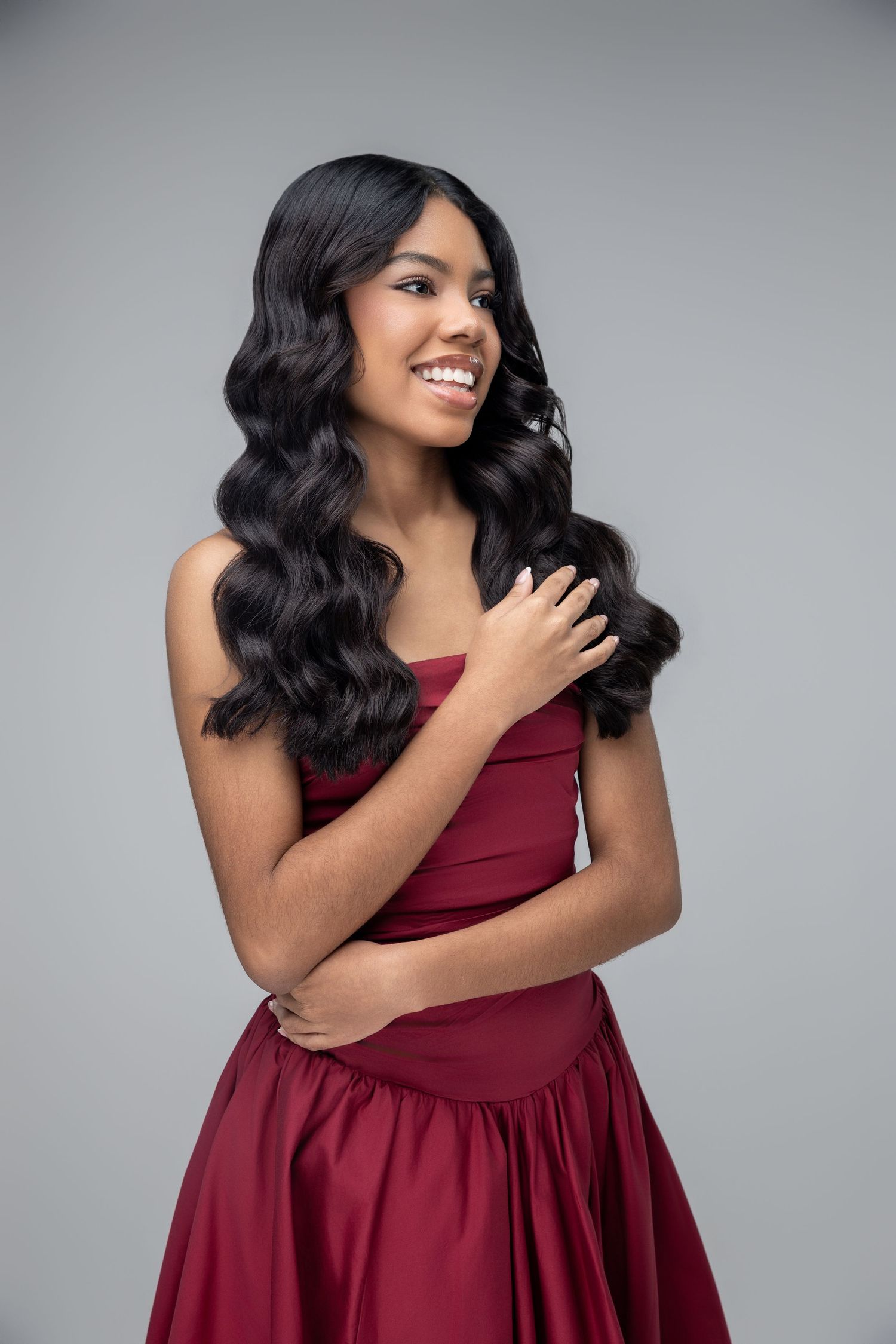 Young woman in a red strapless gown with long wavy black hair, smiling against a gray background.