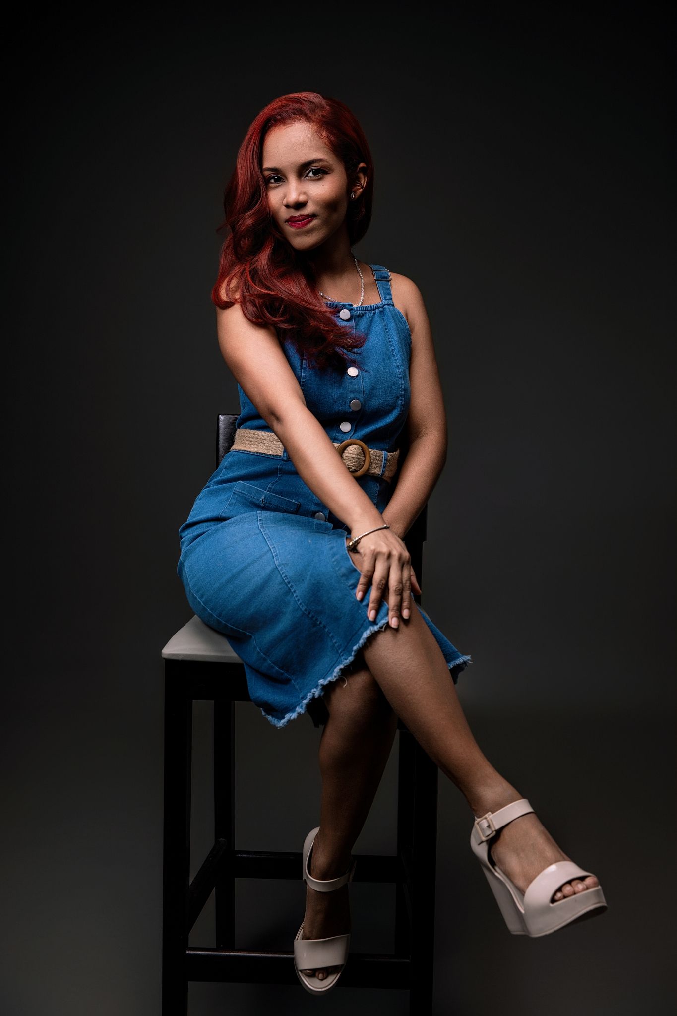 A series of photos showing a person in a blue denim dress and sandals posing on a black chair against a dark background.