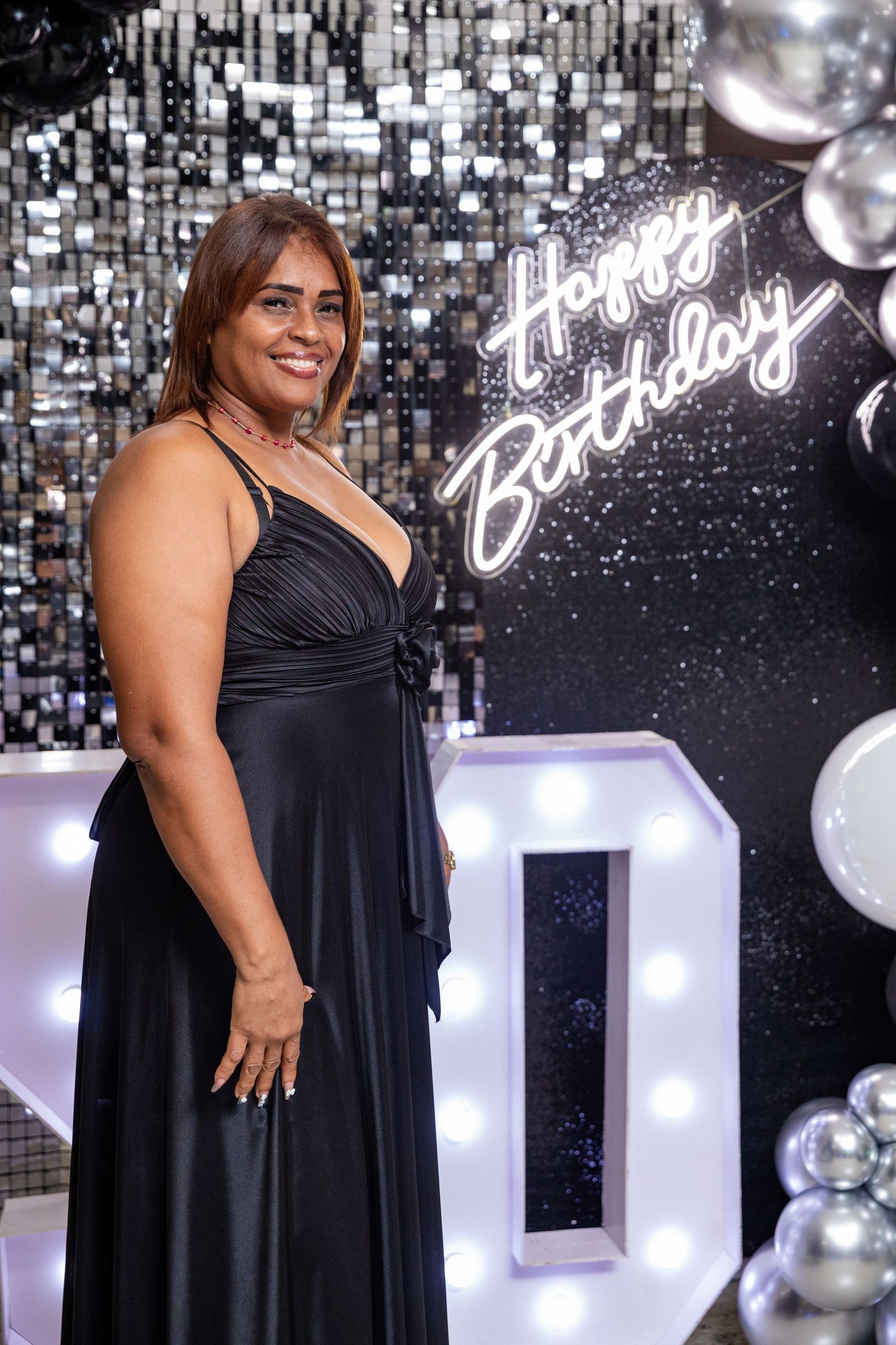 Woman in black gown smiling at a glamorous birthday party with neon Happy Birthday sign and silver balloons.
