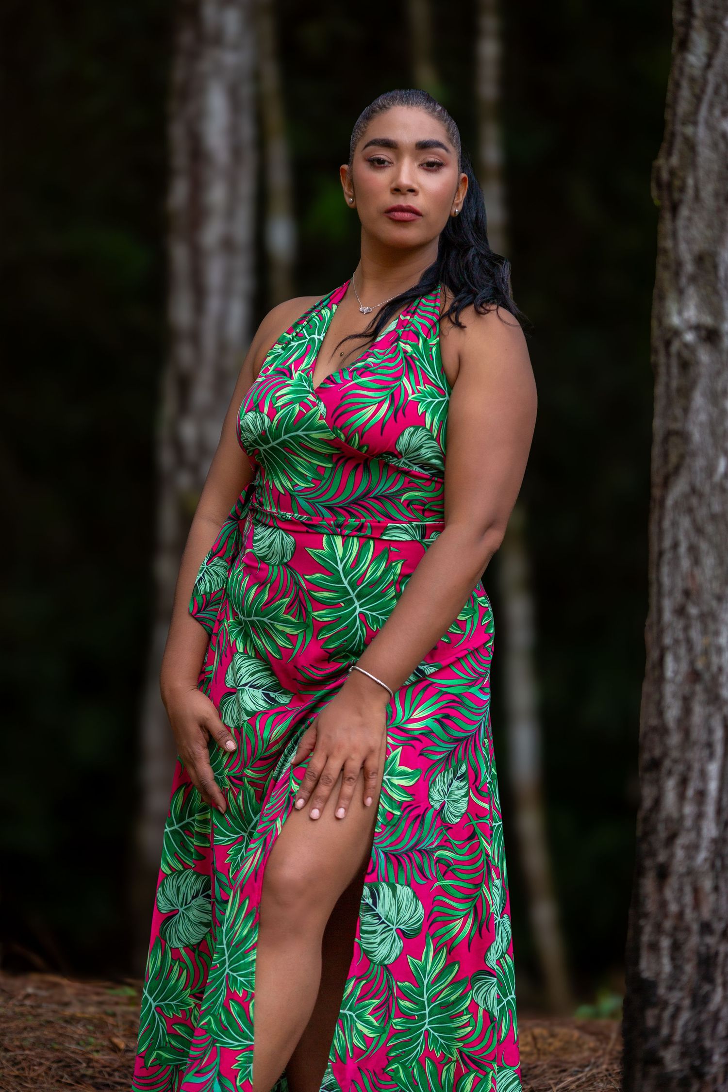 A person in tropical print dress poses confidently among pine trees during outdoor photoshoot.