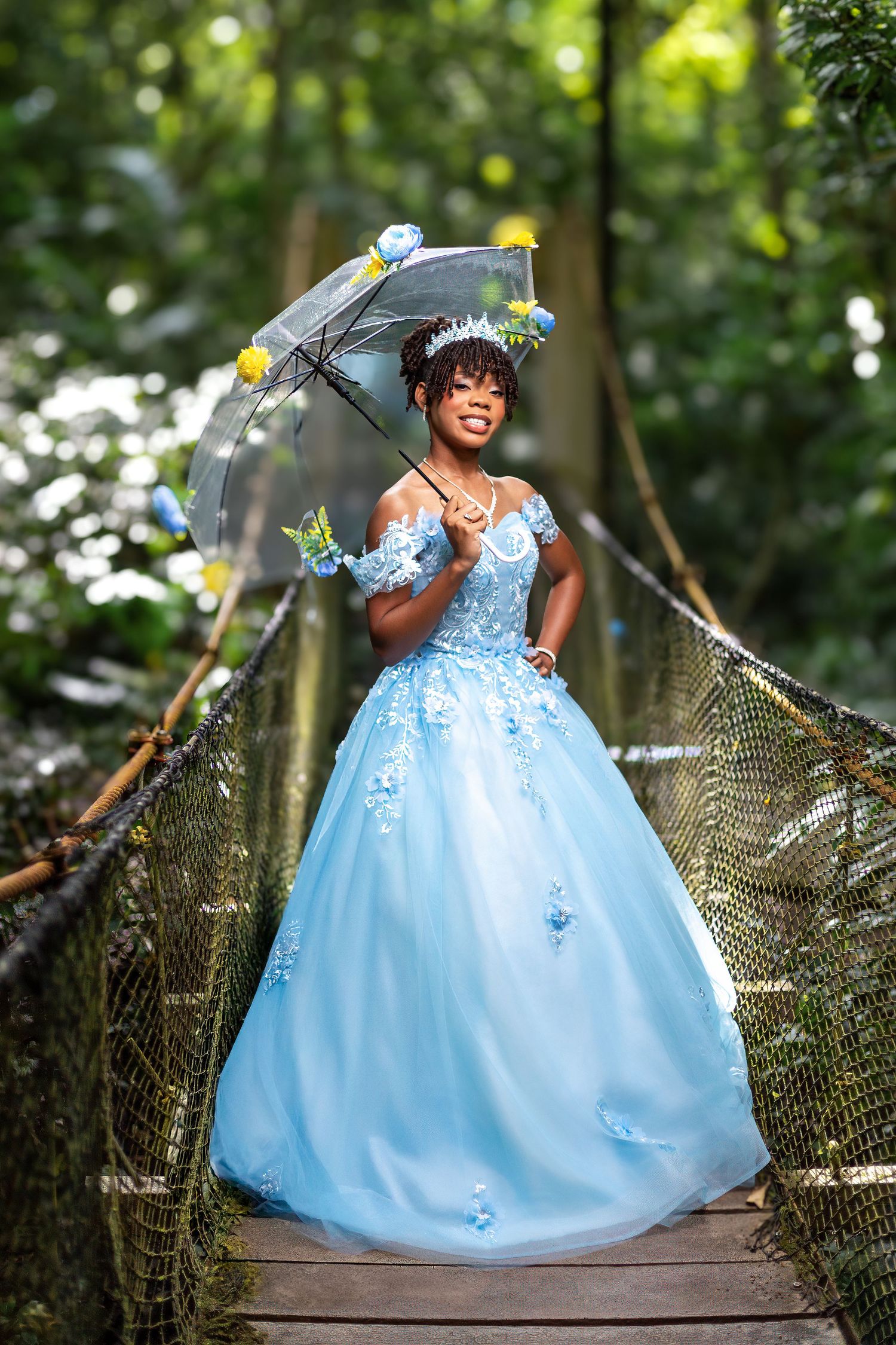 Young woman in light blue ball gown with floral details holds umbrella on wooden bridge in lush green forest.