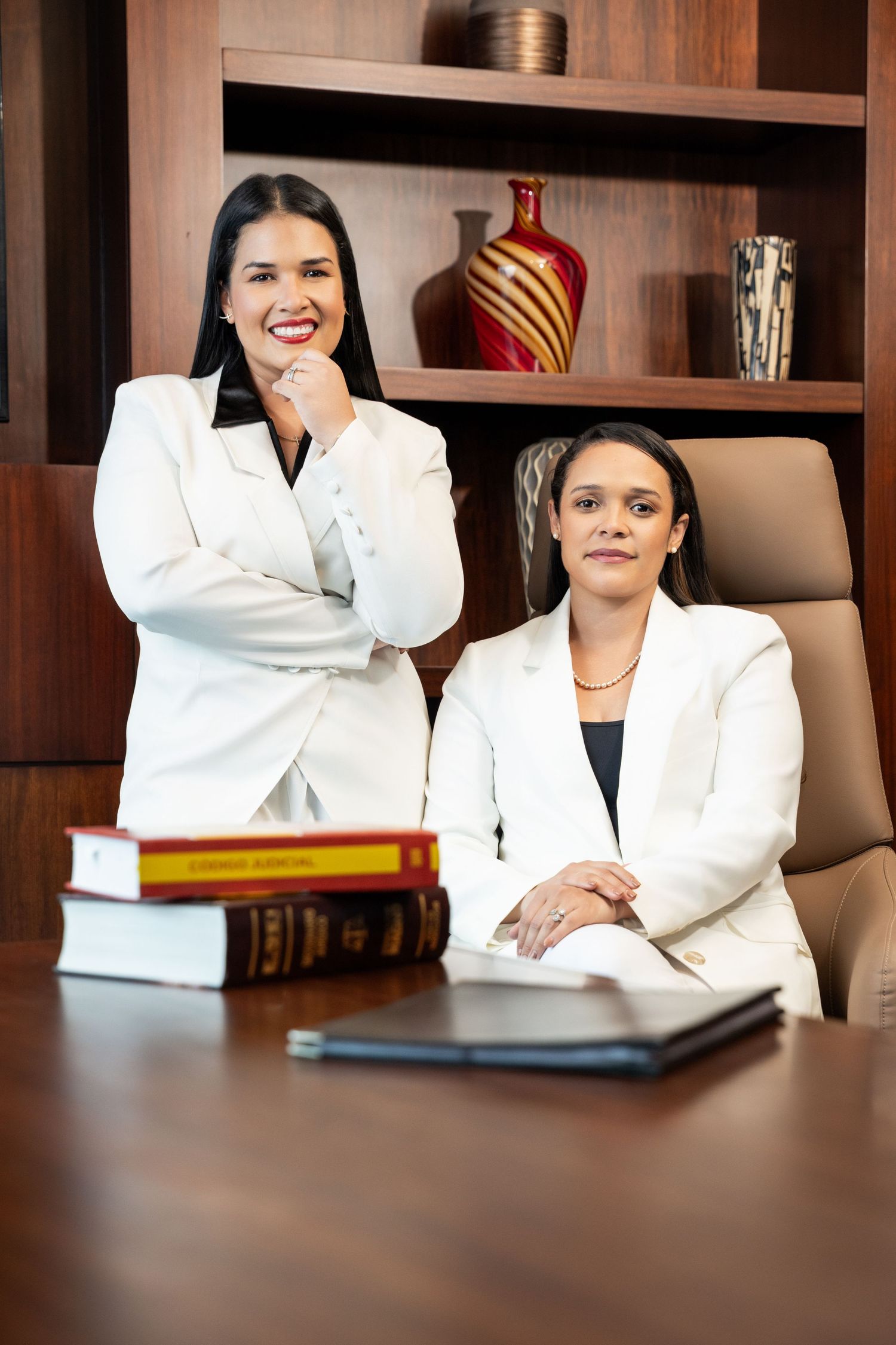 Two professional women in white blazers pose at a wooden conference table with law books nearby.