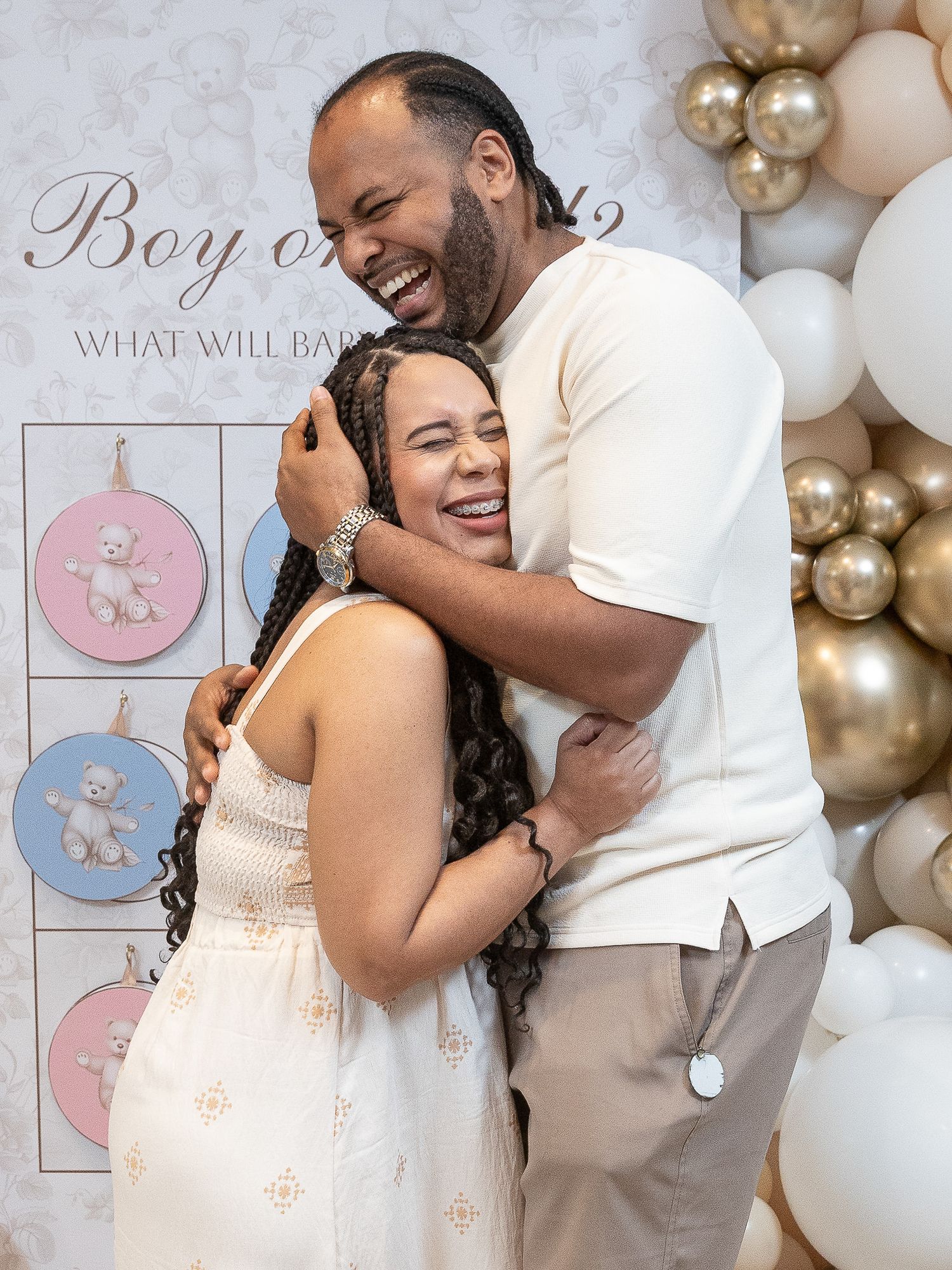 Happy couple embracing and laughing at a gender reveal party with boy or girl balloon backdrop.