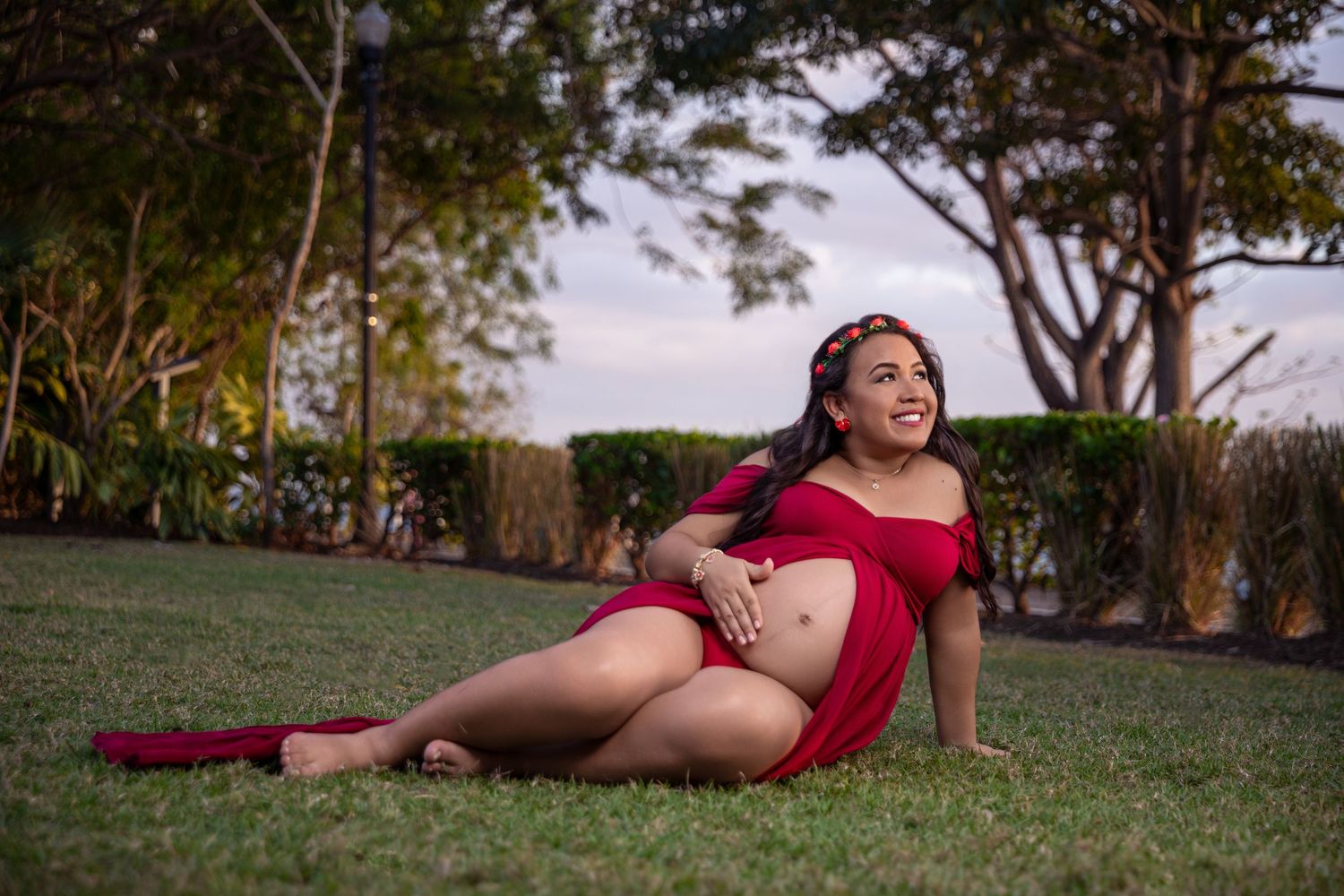 Smiling pregnant woman in red dress sitting on grass in a park during a maternity photo shoot.