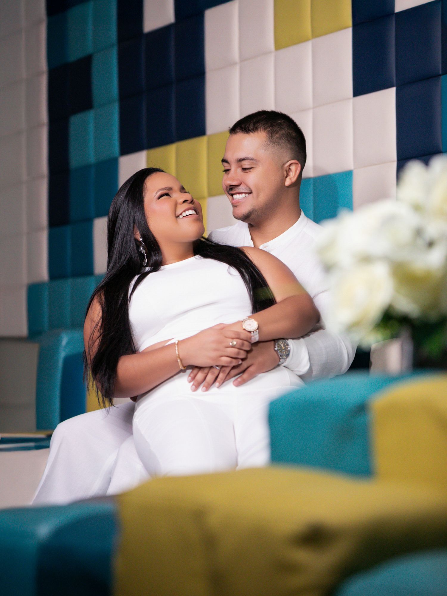 Happy couple in white outfits embracing and smiling in a colorful modern lounge setting.