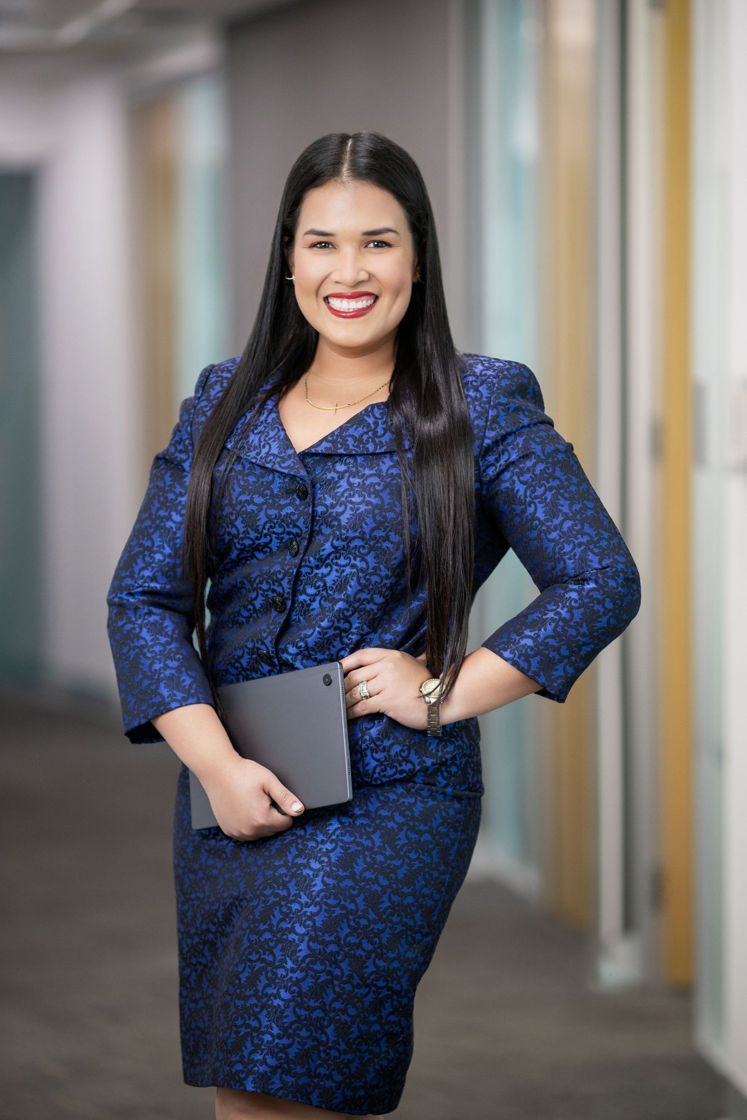 Professional businesswoman in blue patterned suit holding tablet, smiling confidently in modern office hallway.