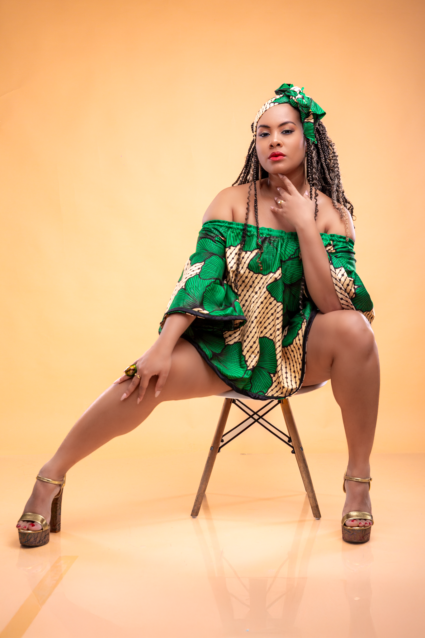 Model poses on chair wearing green and gold patterned dress against peach-colored backdrop.