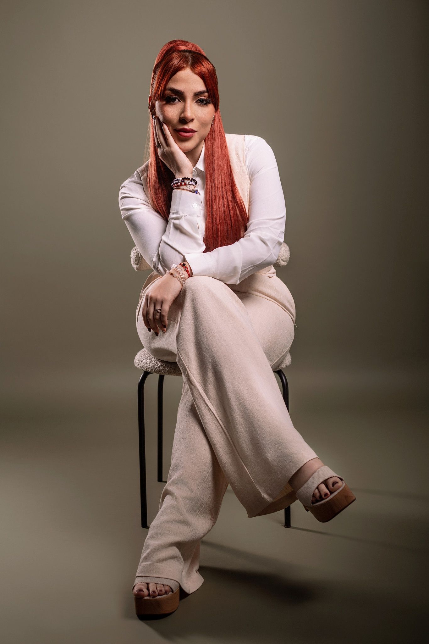 Model with long red hair wearing white outfit poses in various seated positions against gray backdrop for fashion shoot.