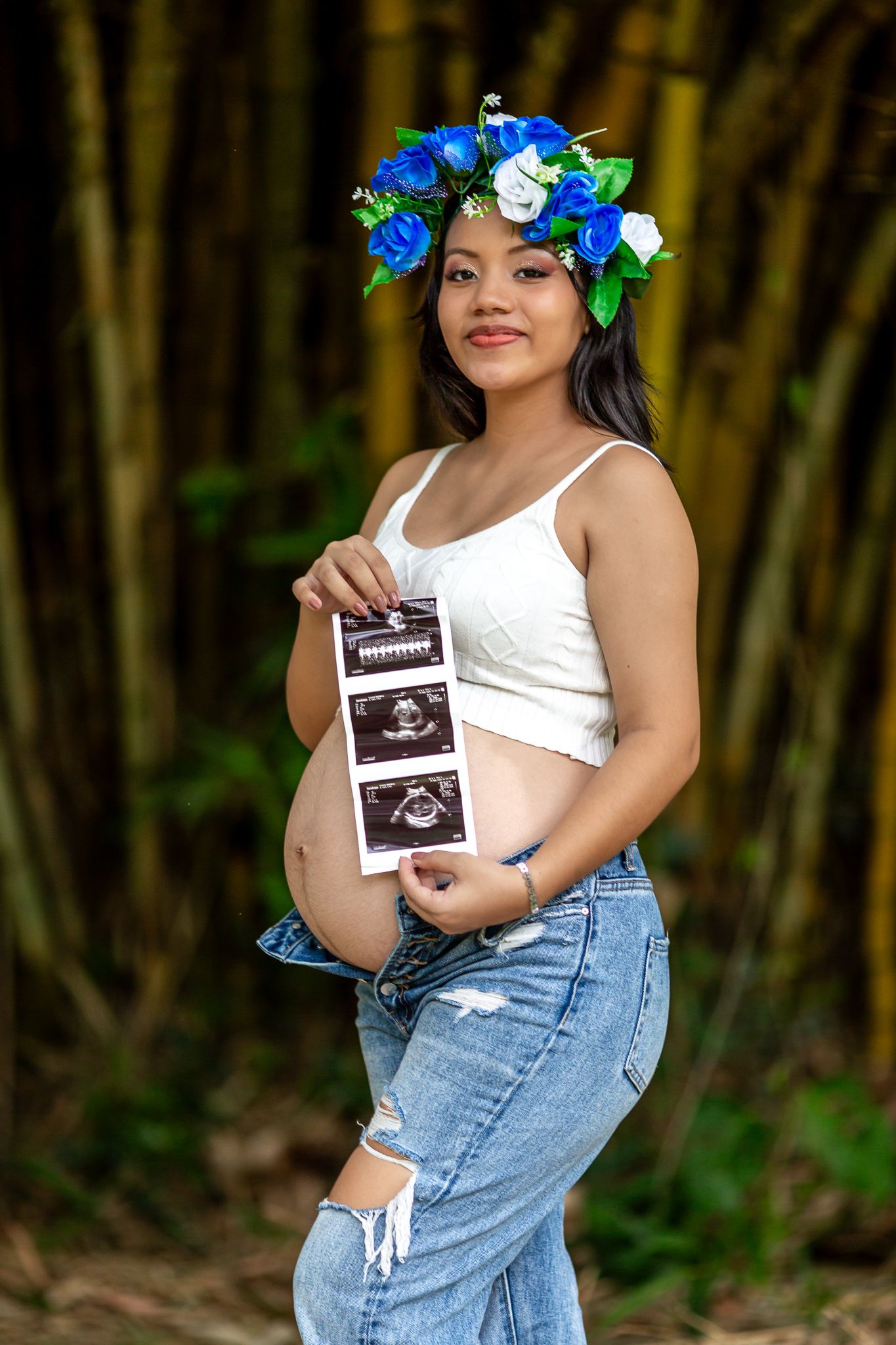 Pregnant woman in distressed jeans holds ultrasound photos while wearing blue floral crown.