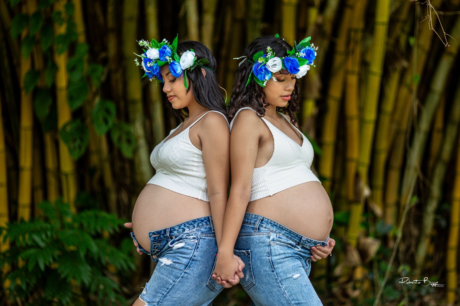 Two pregnant women in matching floral headpieces and denim jeans pose back-to-back in front of bamboo stalks.
