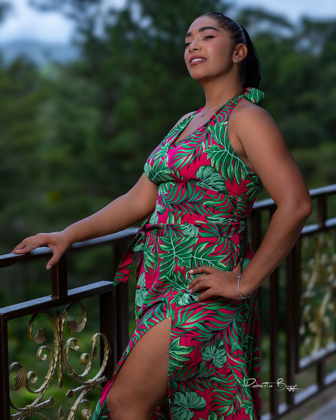 A woman poses in a vibrant red and green tropical print dress with a side slit while leaning against an outdoor railing.