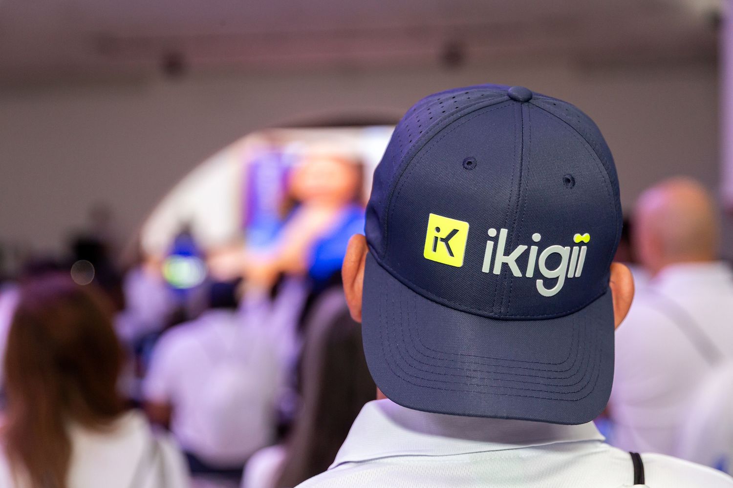 A person wearing a navy blue hat with the Ikigi logo on the front, standing in a crowd of people.