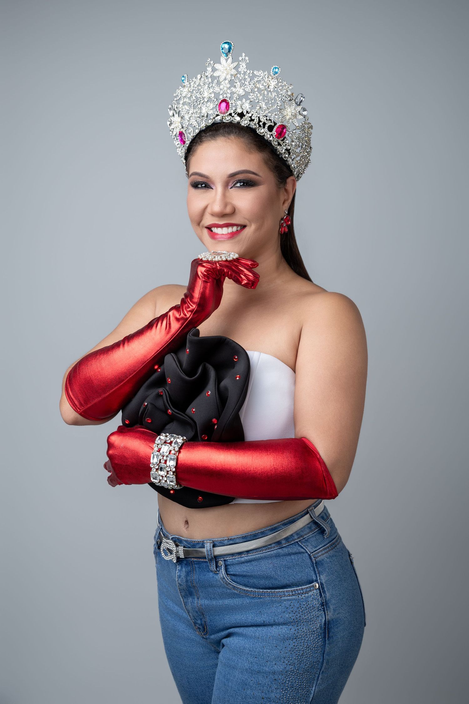Woman wearing a jeweled crown, red satin gloves, white top, and jeans poses confidently against a gray background.