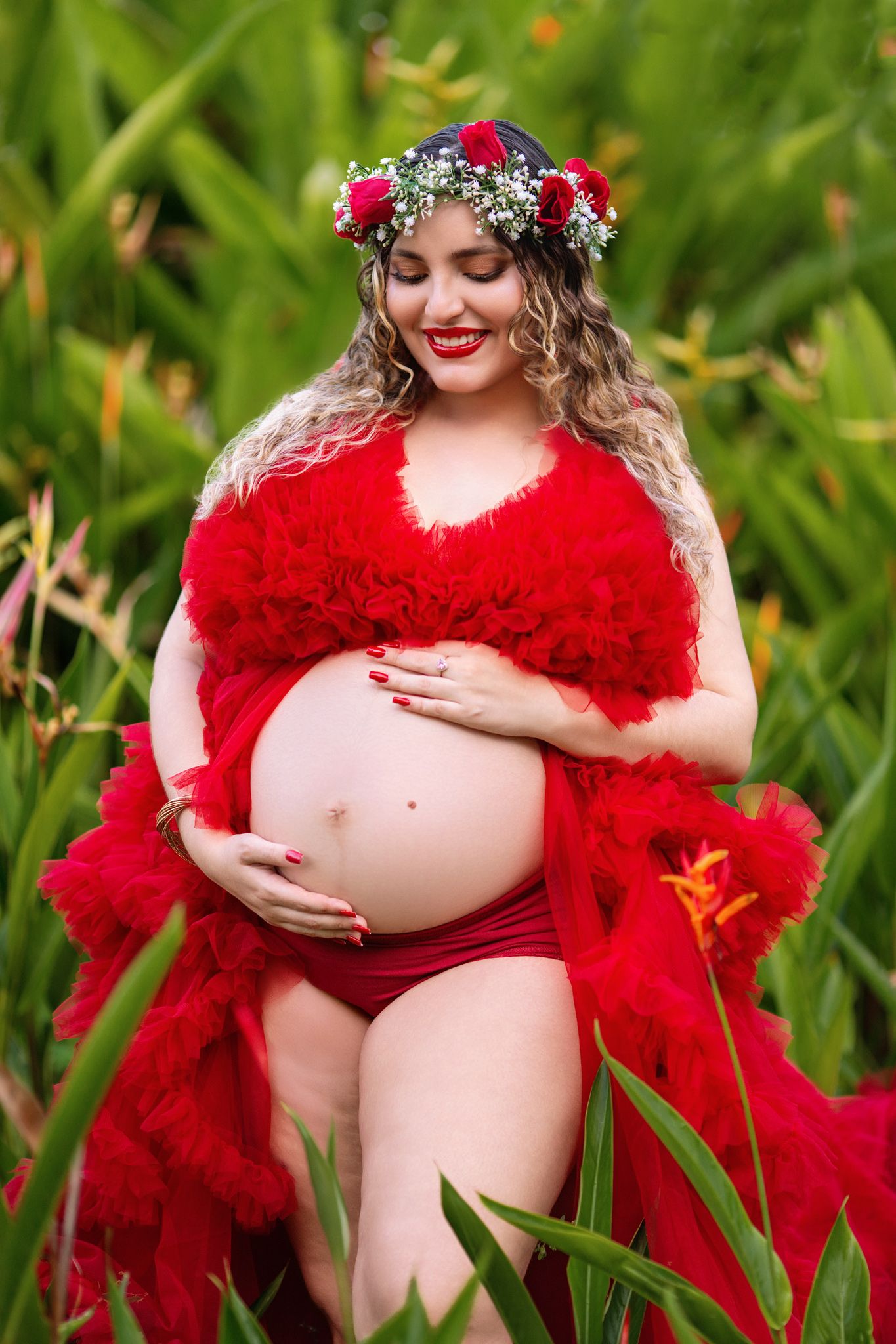 A maternity photoshoot in nature features vibrant red tulle fabric against lush green foliage.