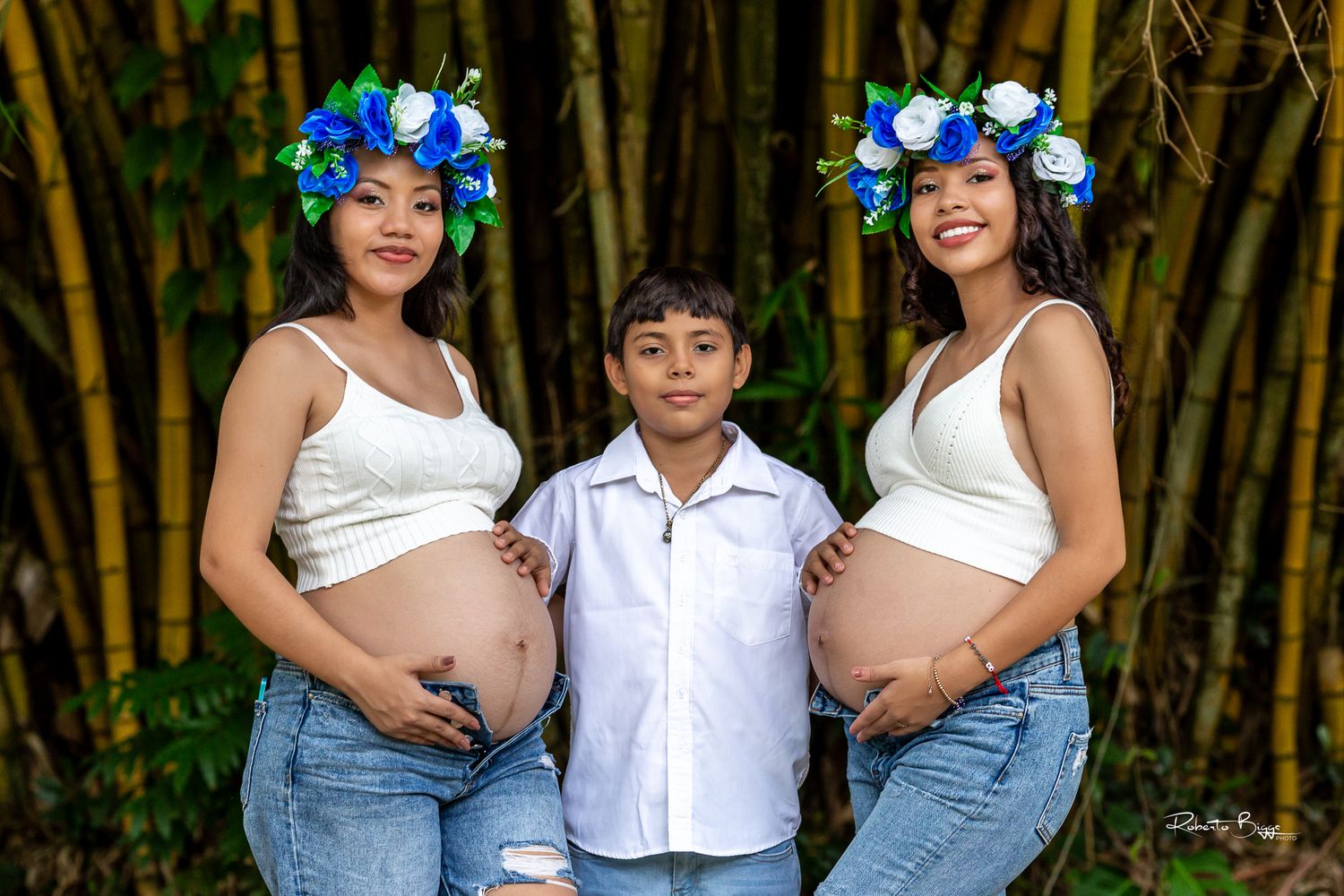 Three people pose outdoors by bamboo trees, with matching blue floral crowns and exposed baby bumps in the photo.
