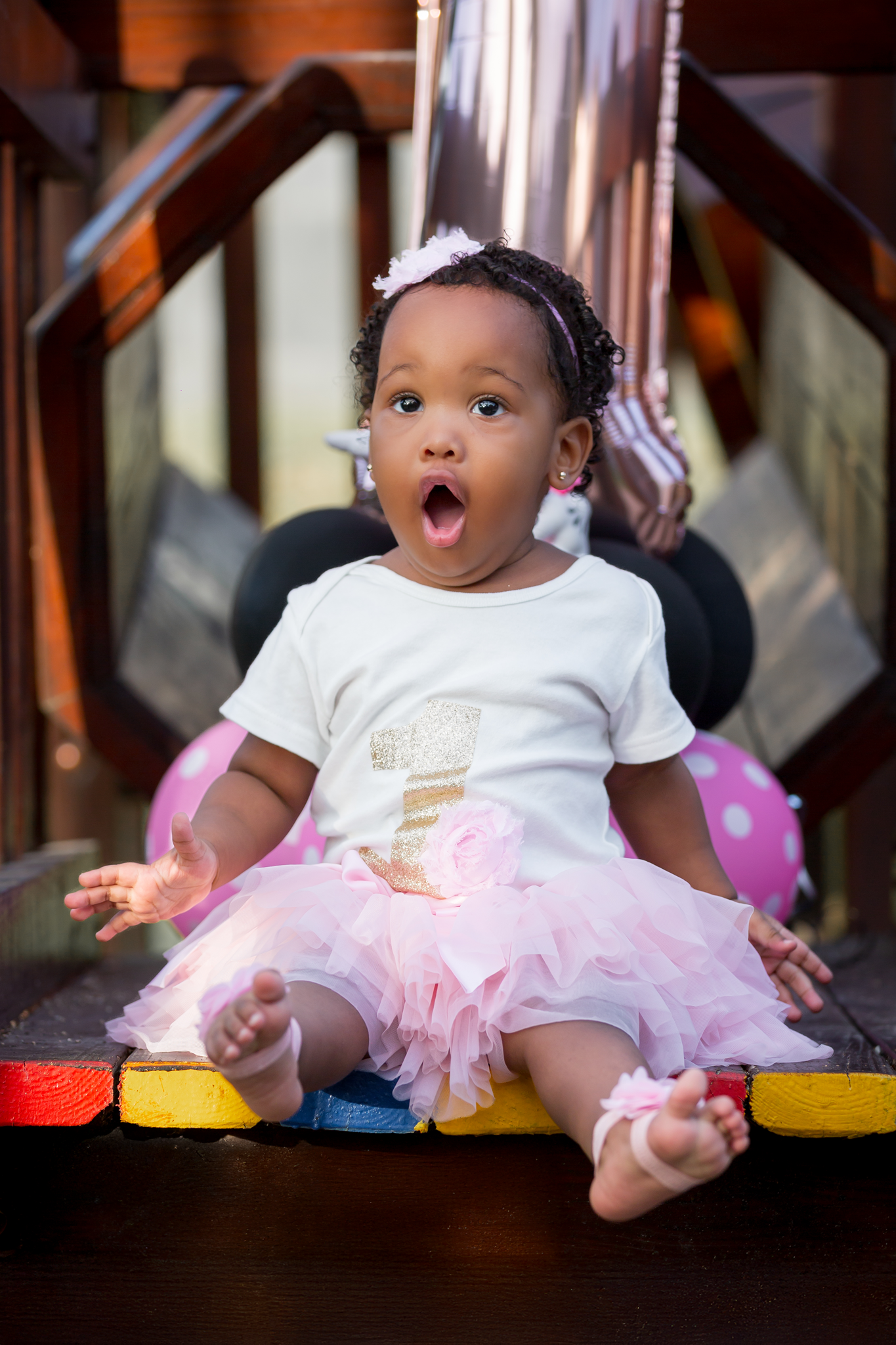 Baby in a white shirt and pink tutu skirt sits on colorful steps with an excited expression.