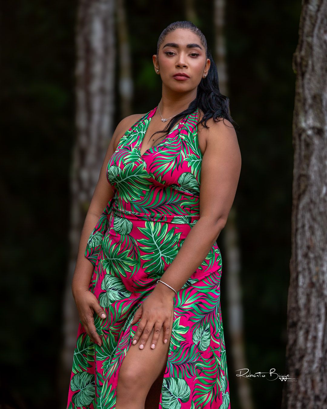 A graceful figure poses in a vibrant tropical print dress with green monstera leaves and pink flowers against a forest backdrop.