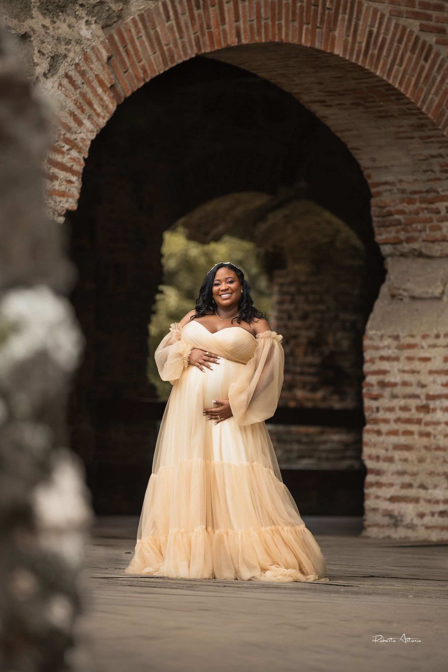 Pregnant woman in elegant champagne tulle gown poses under brick arch for maternity photo session.