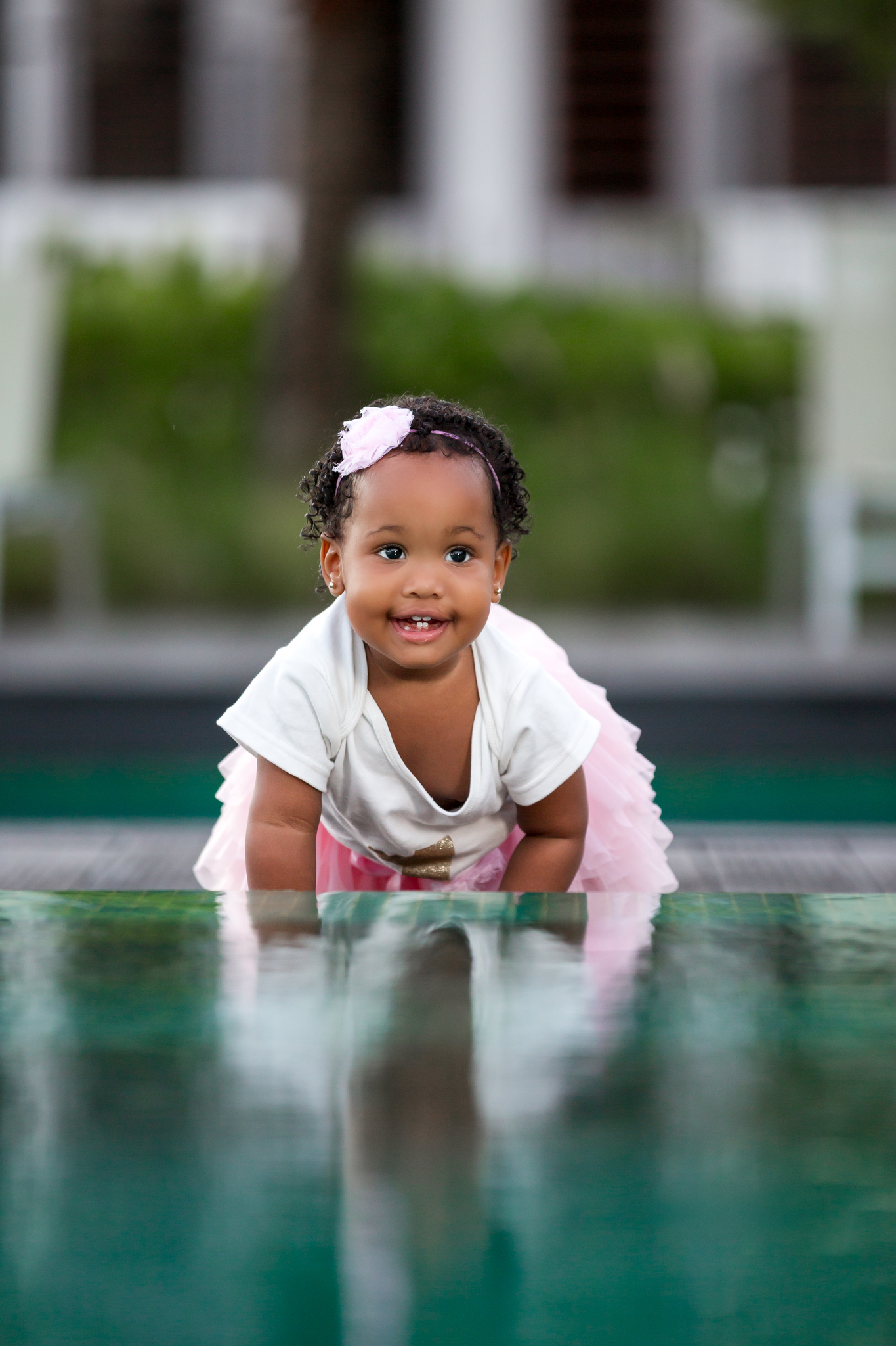 A baby in white dress crawling near a reflective surface with a bright smile.