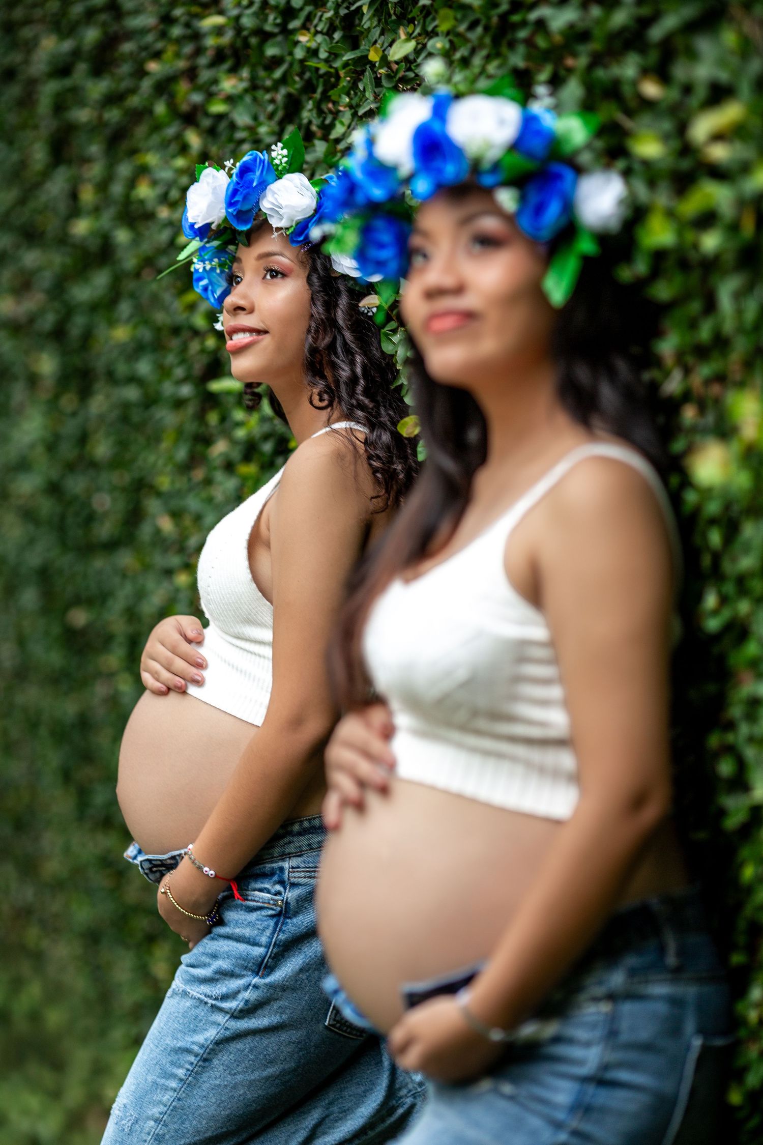 Two pregnant women in white tops and jeans pose against a green hedge wearing matching blue floral crowns.