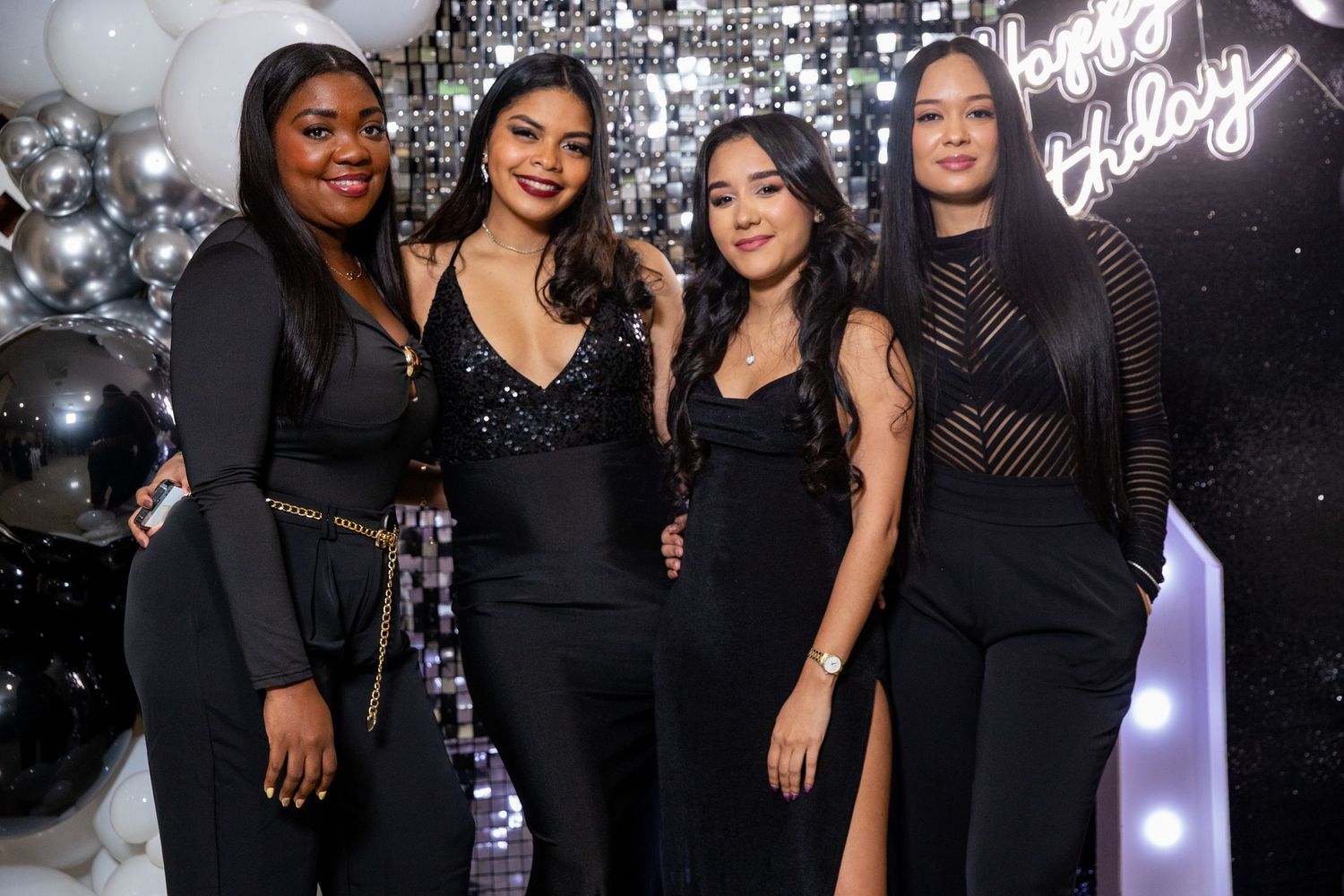 Four women in elegant black outfits pose together at a birthday party with silver balloon decorations.