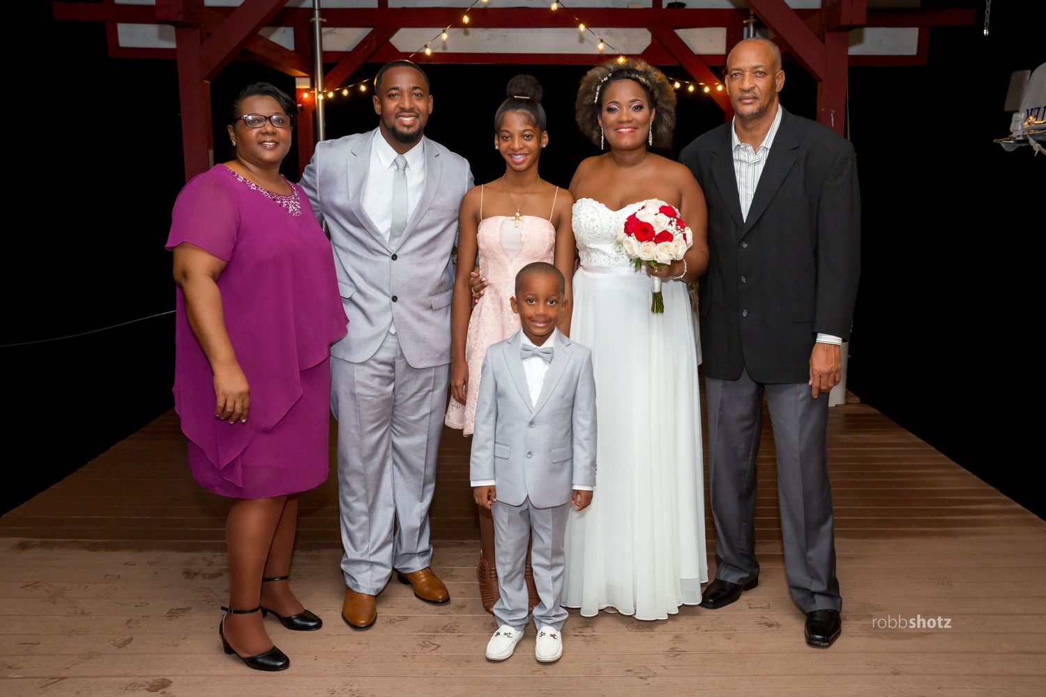 Wedding party group photo with formal attire on wooden deck at night.