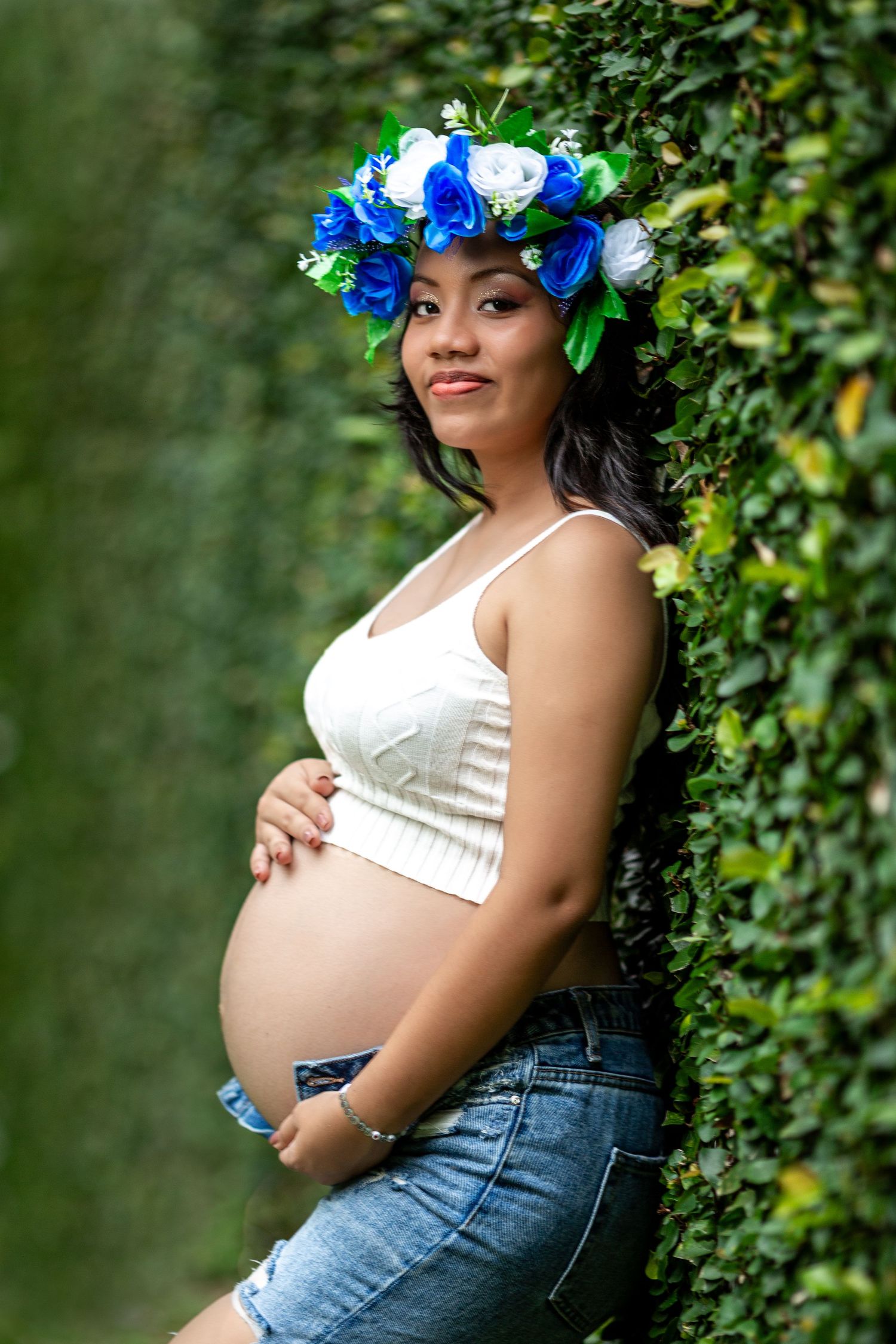 A maternity photo shoot in a garden setting shows a figure wearing a white top, blue flower crown and denim against greenery.