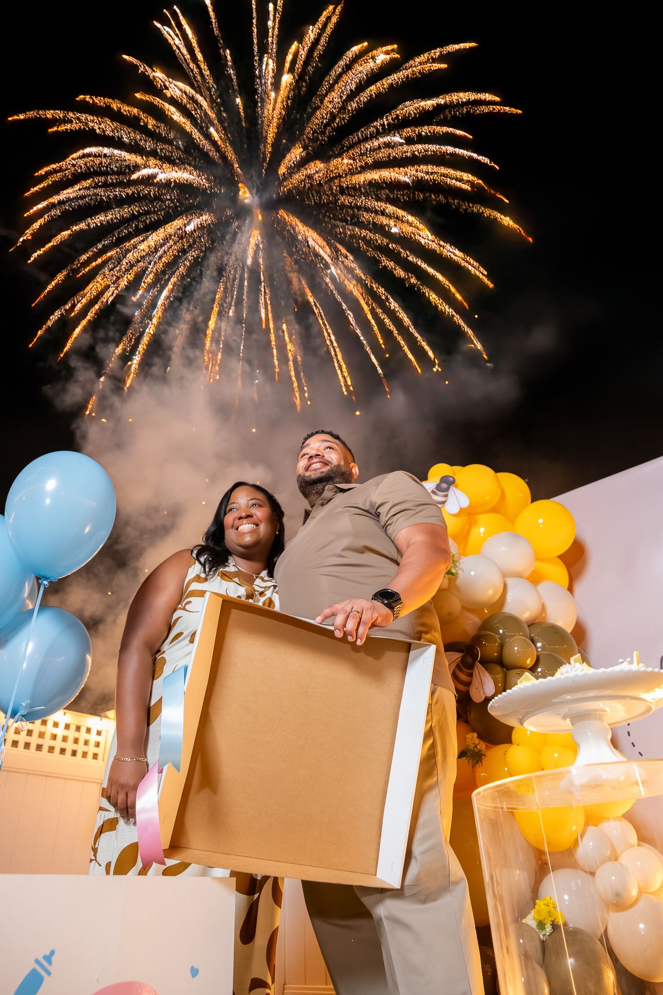 Couple holding a box at a gender reveal party with blue and yellow balloons and colorful fireworks lighting the night sky.