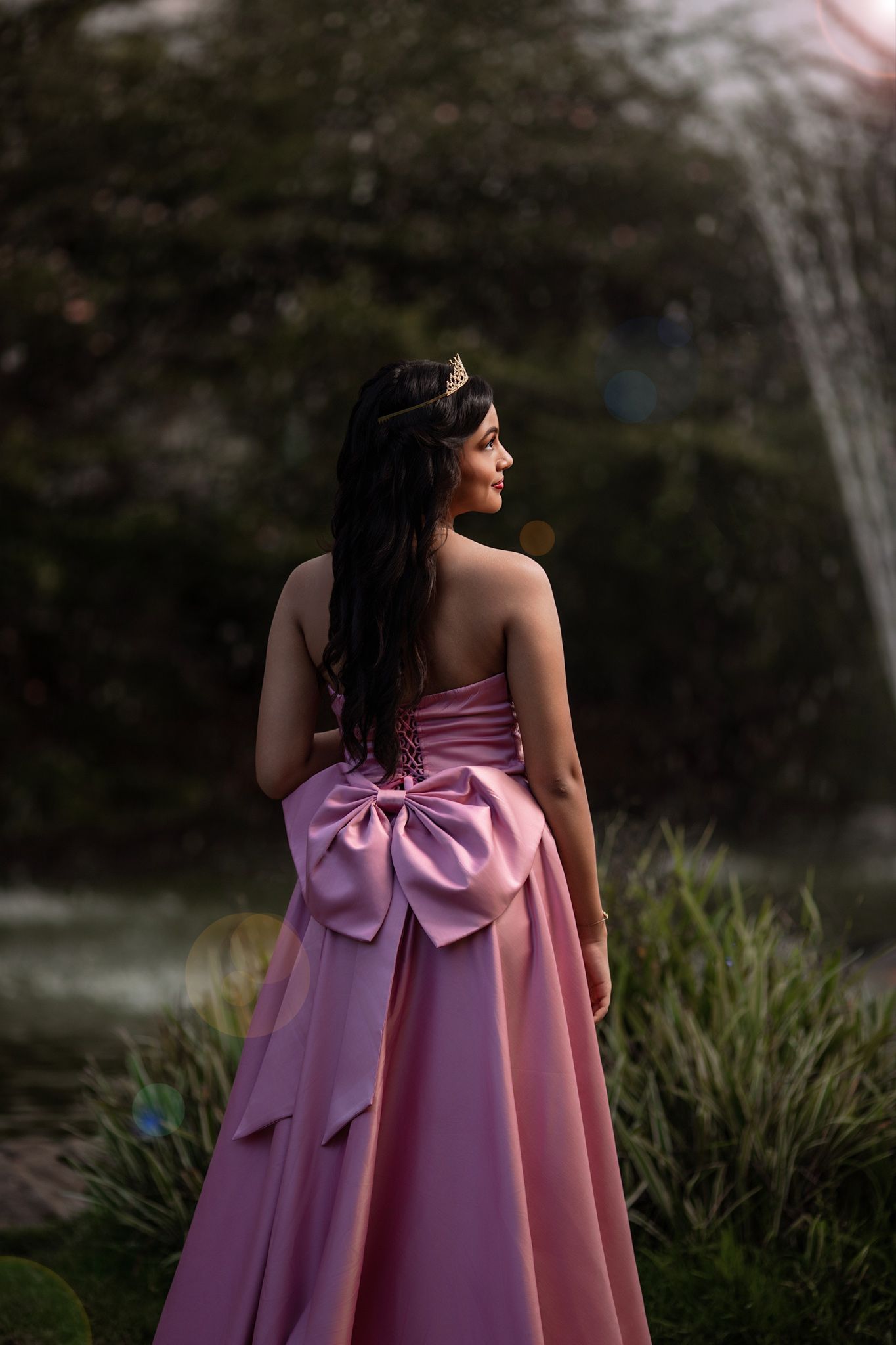 A woman in a pink formal gown with a large bow detail stands in a garden at dusk.