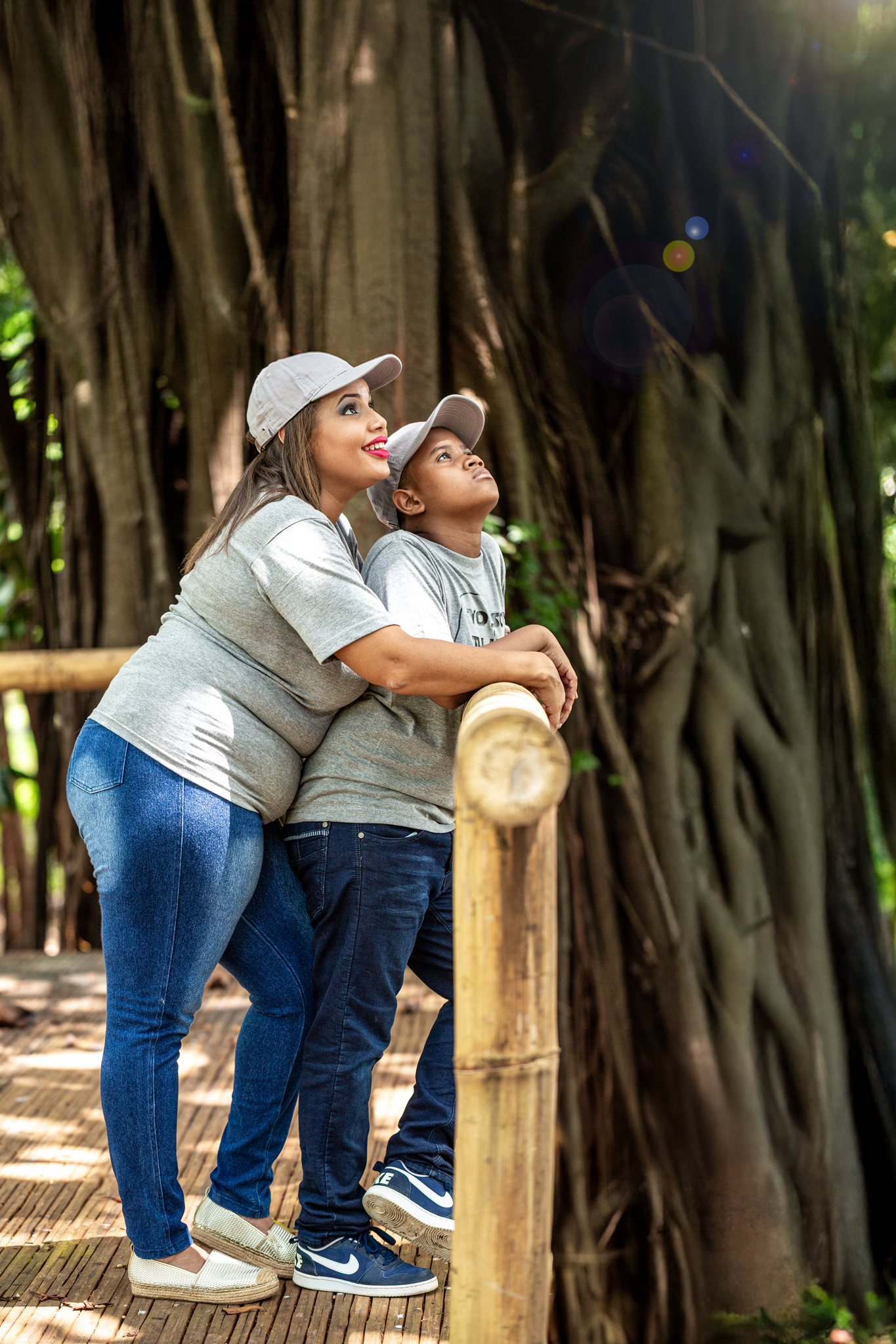 Two people in white baseball caps and casual clothing share a joyful moment together while looking up at trees in a park.