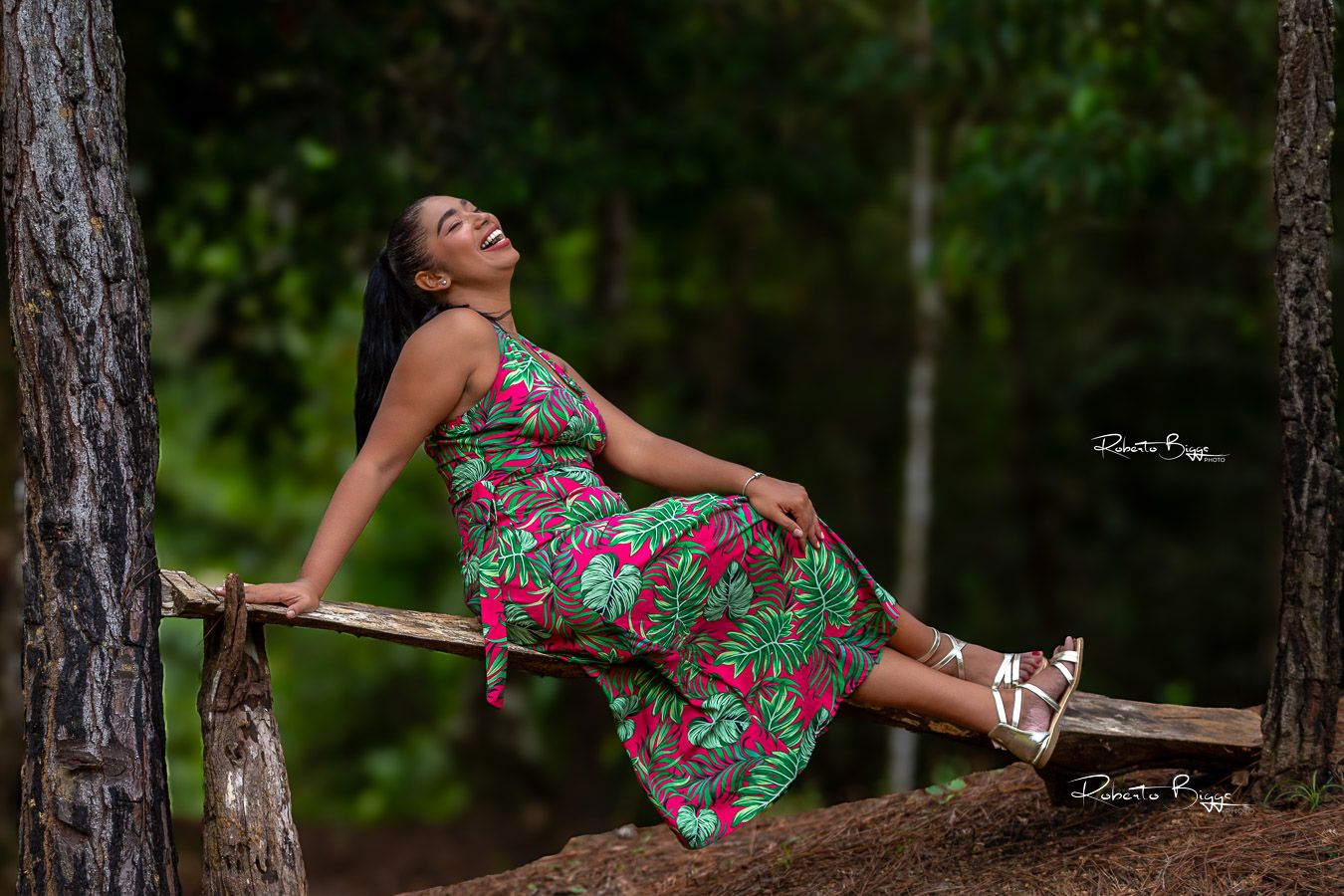 A person in a vibrant floral dress reclines on a rustic wooden bench between tree trunks in a forest setting.