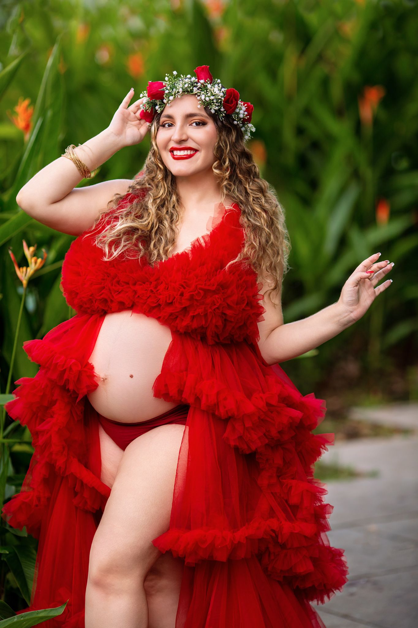 Maternity photoshoot featuring flowing red tulle dress and floral crown in outdoor garden setting.