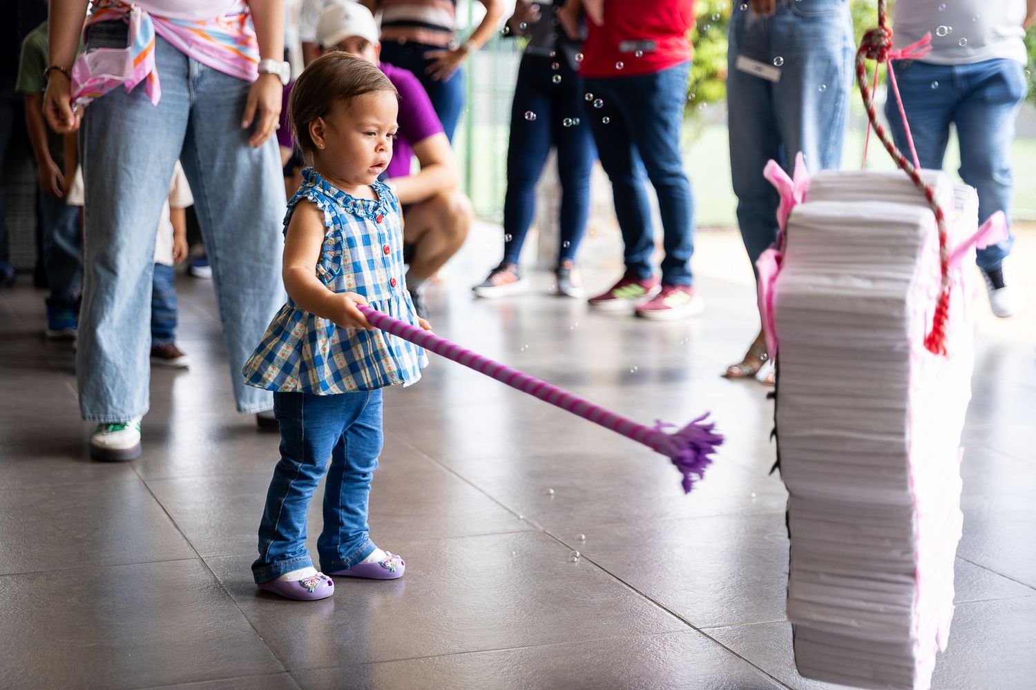 Young girl in plaid dress swings pink stick at a pinata during a birthday party celebration indoors.