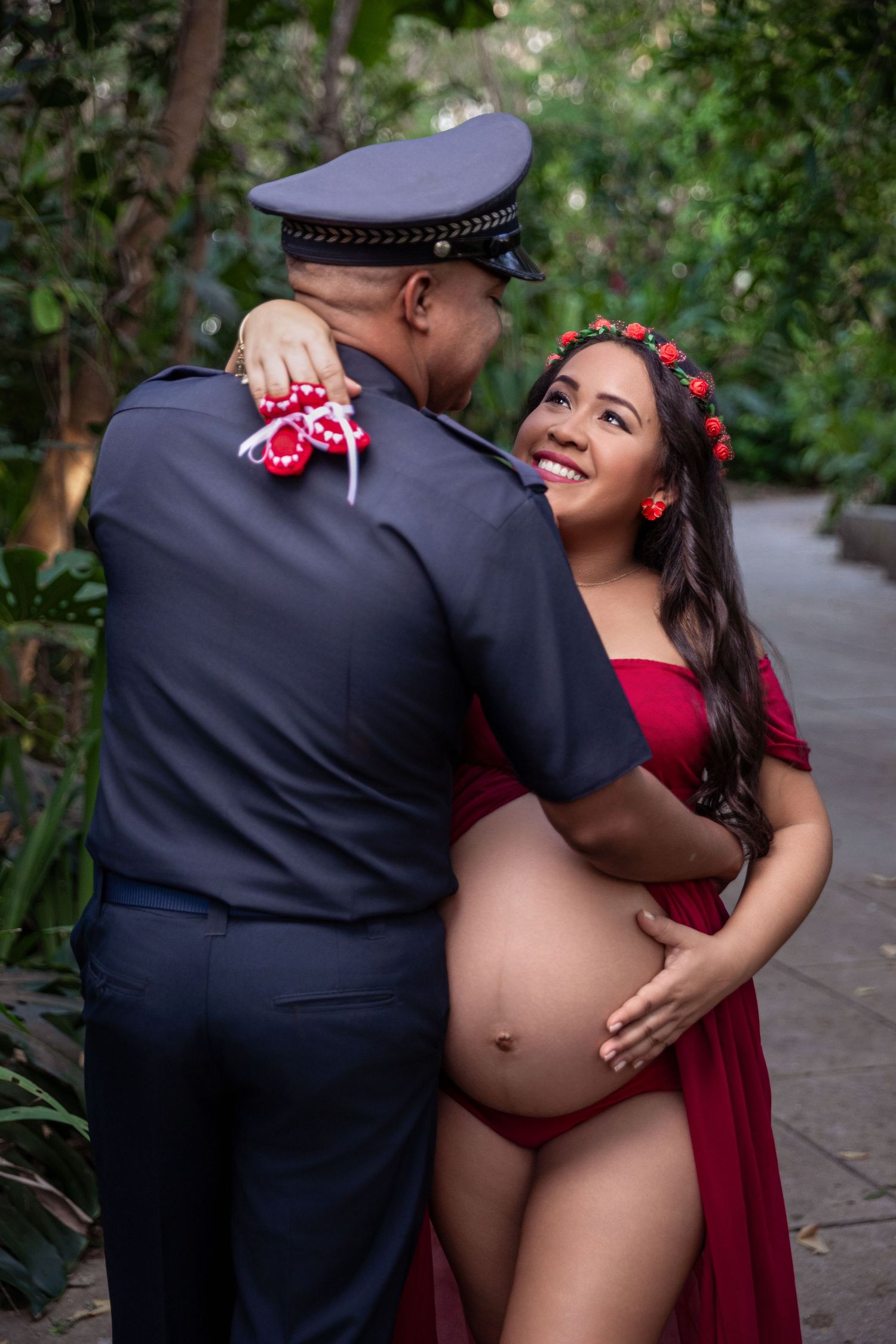 Pregnant woman in red dress with flower crown embraces a man in police uniform holding tiny baby shoes outdoors.