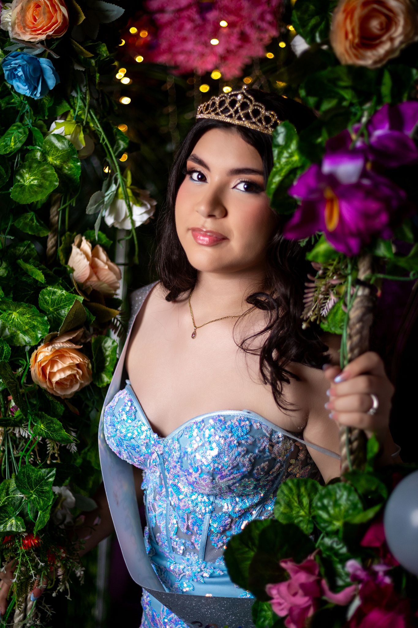 A dreamy portrait in a blue gown surrounded by pink and orange roses and twinkling lights in a garden setting.