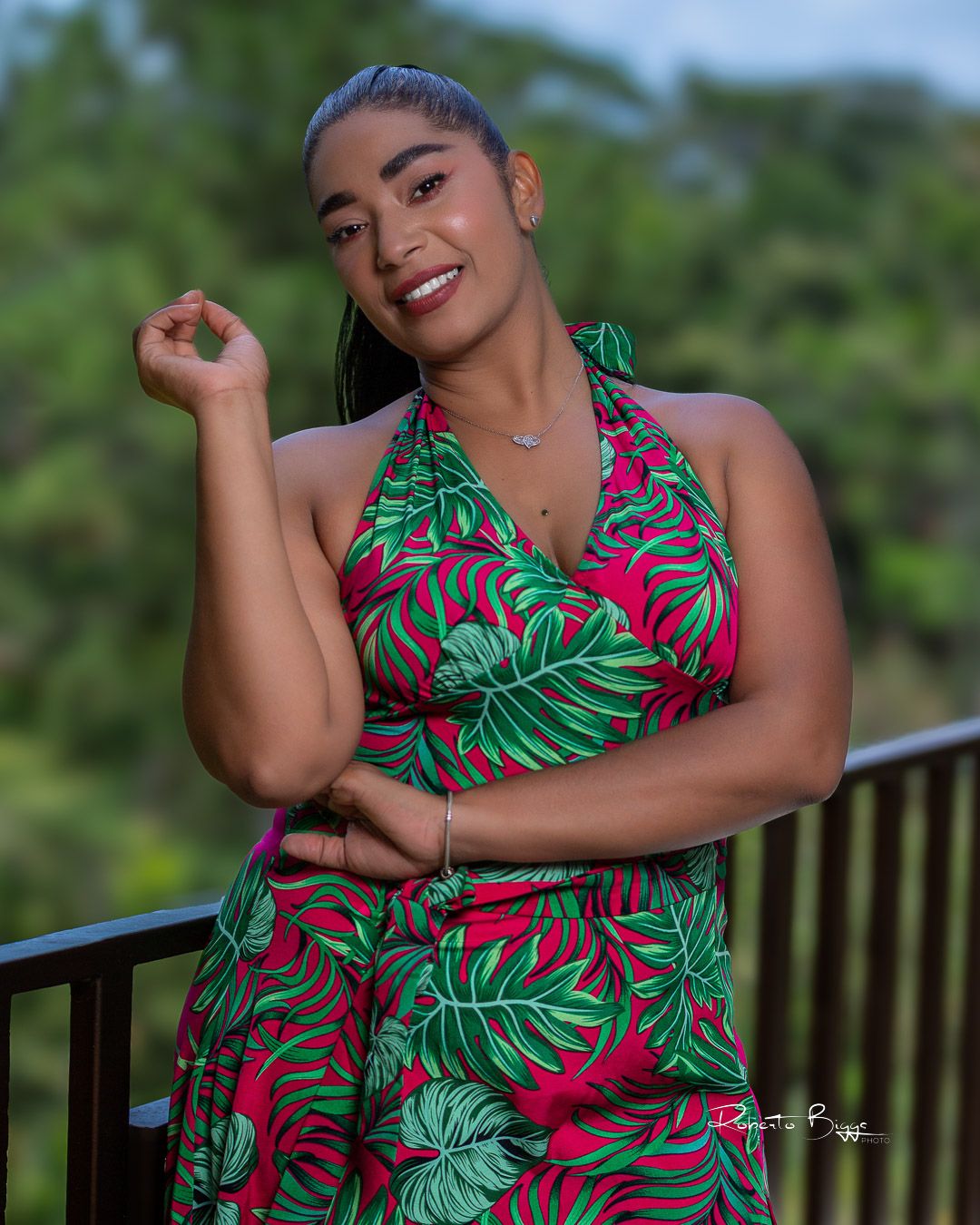 Person models tropical print dress in red and green palm leaf pattern while posing on outdoor deck.
