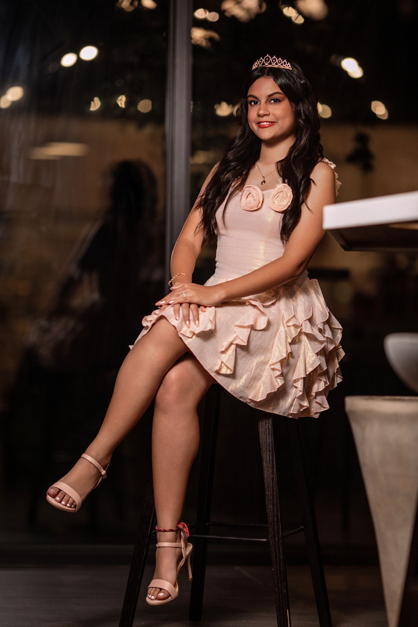 A person in a pink dress and tiara sits on a dark wooden stool in a dimly lit setting.