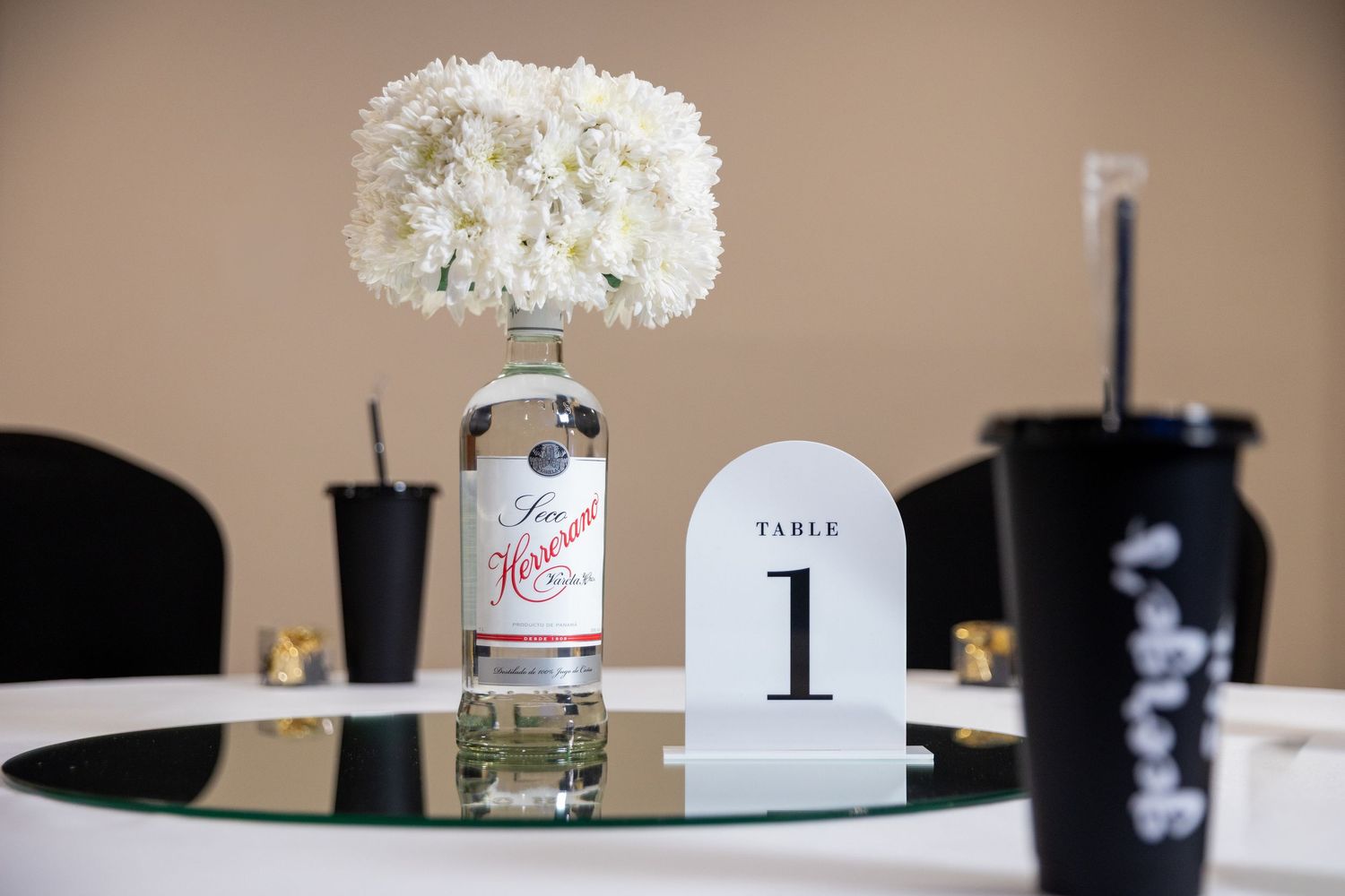 Wedding table centerpiece with white flowers in a liquor bottle, table number one sign, and black cups on a glass table.