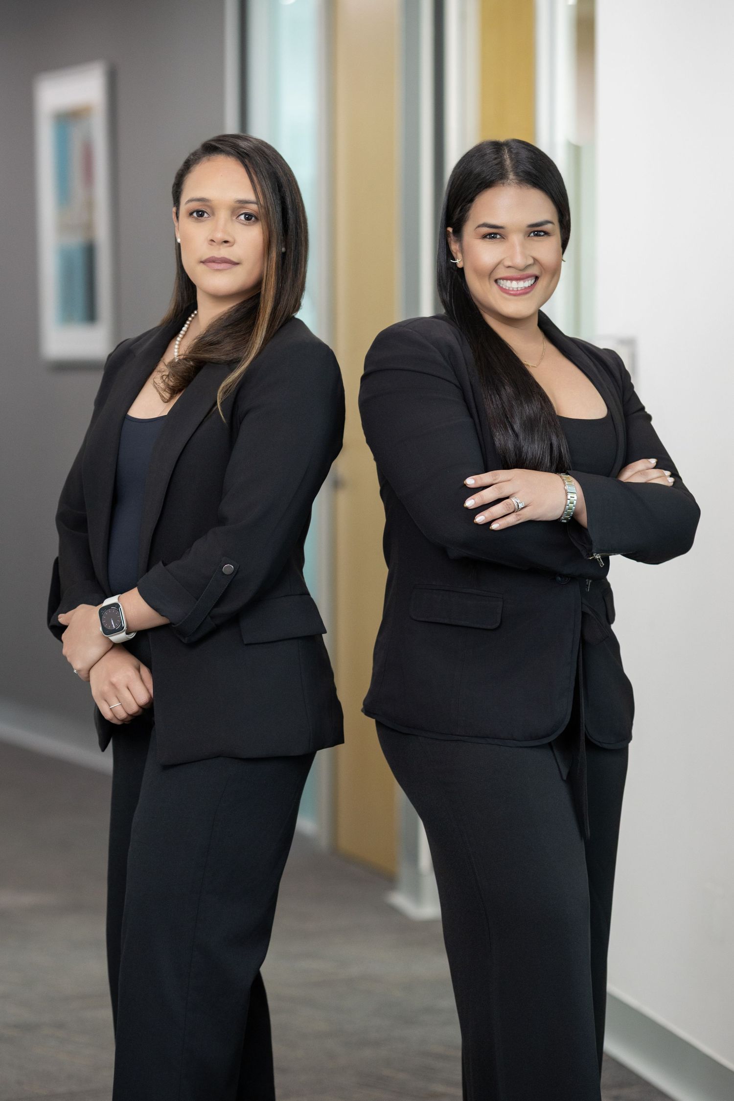 Two professional women in black business suits standing back-to-back in a modern office hallway.