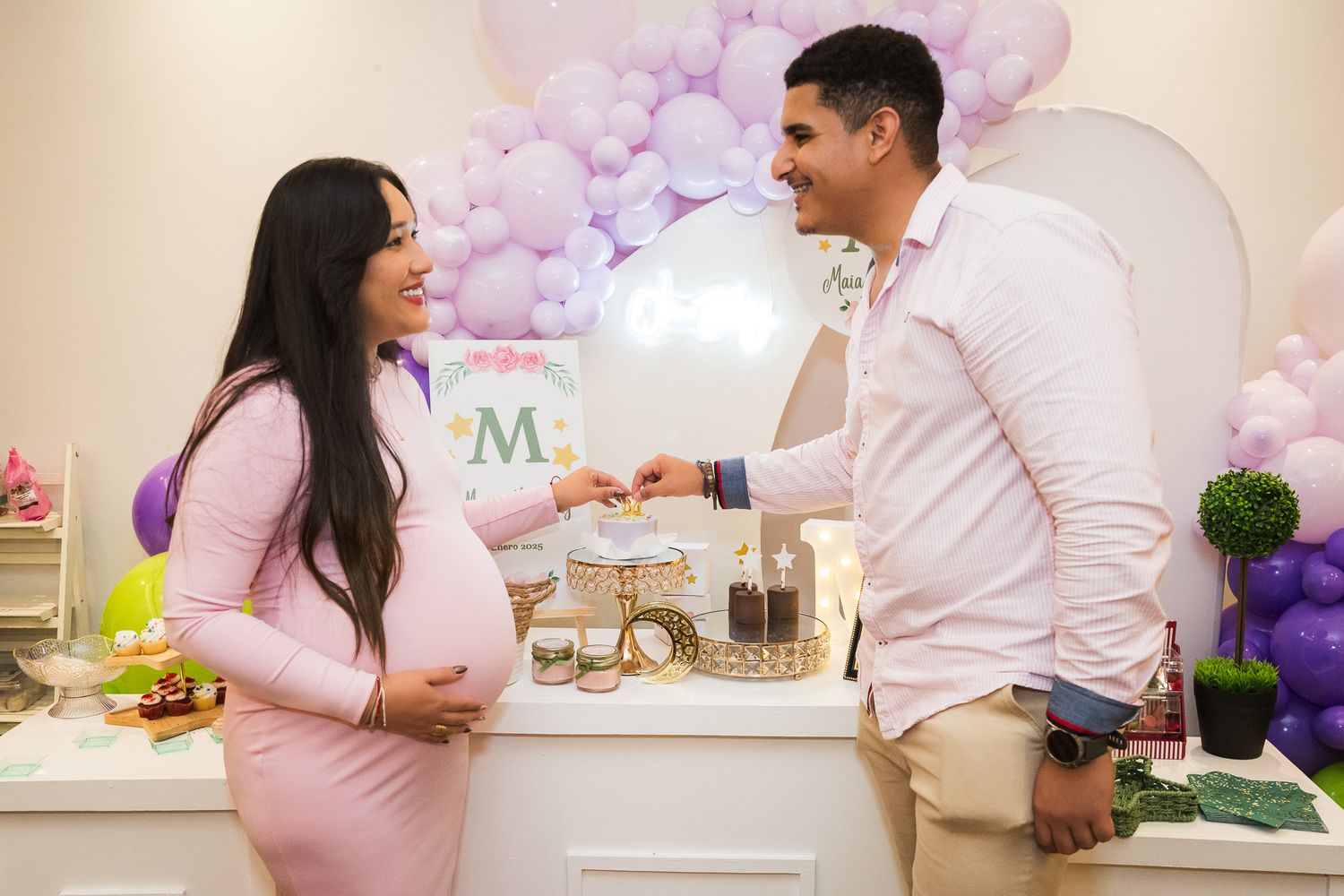 A couple celebrates at a baby shower with pink and purple balloon decorations and party treats on display.