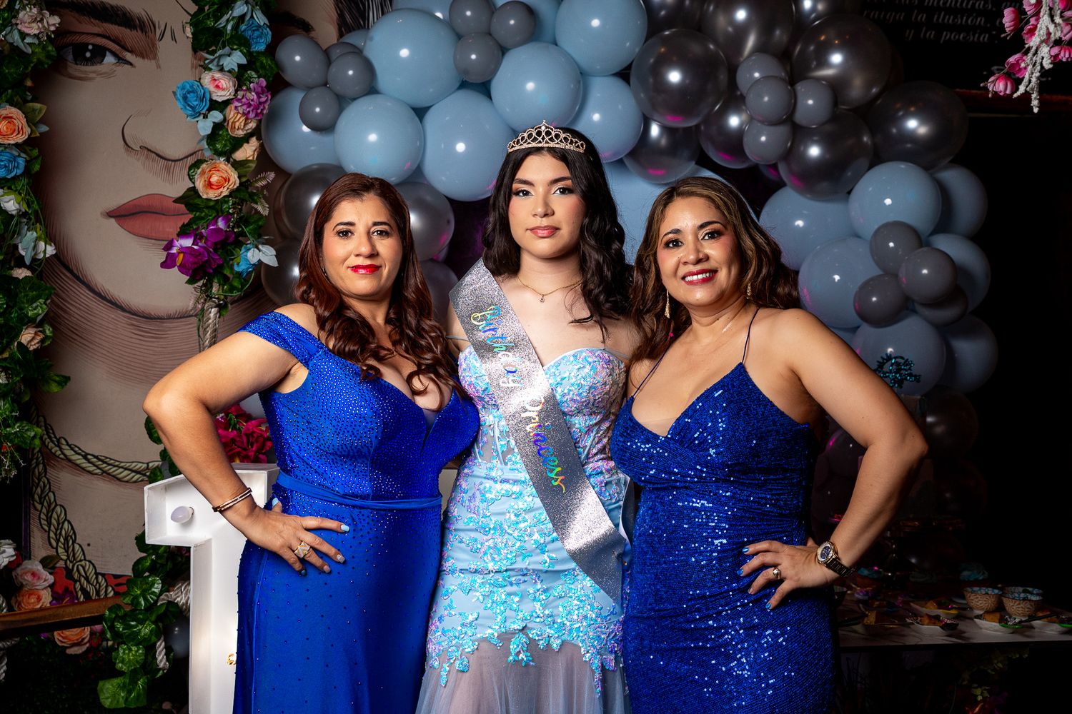 Three people in formal evening attire pose together in front of a balloon arch and decorative backdrop.