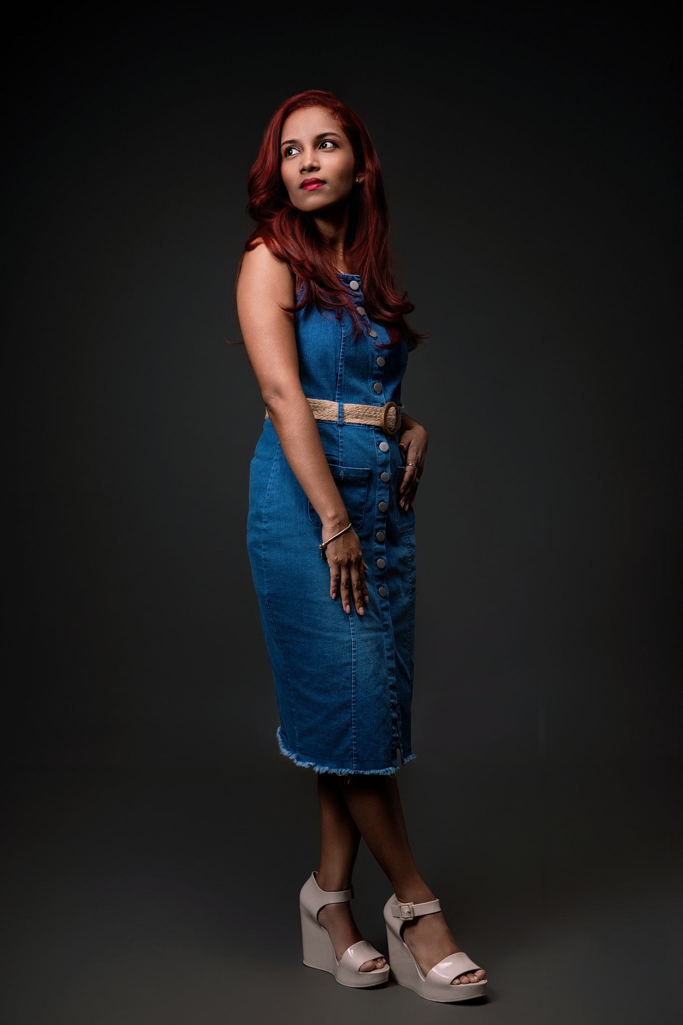Person in blue denim overall dress and platform sandals poses against dark backdrop in fashion photoshoot series.
